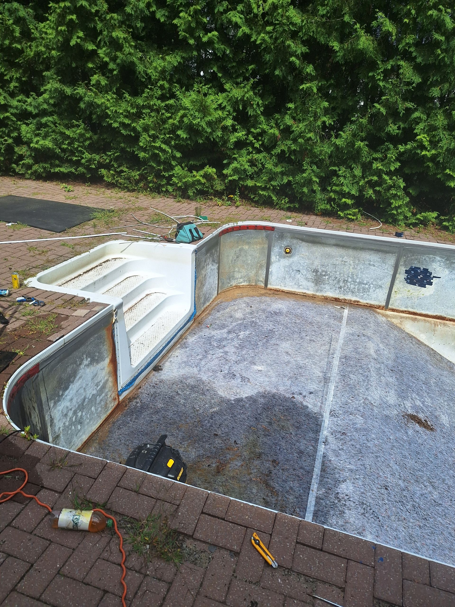 Une piscine en construction dans un jardin, encore vide, laisse apparaître des parois en acier, des marches blanches et un fond en matériau grossier.