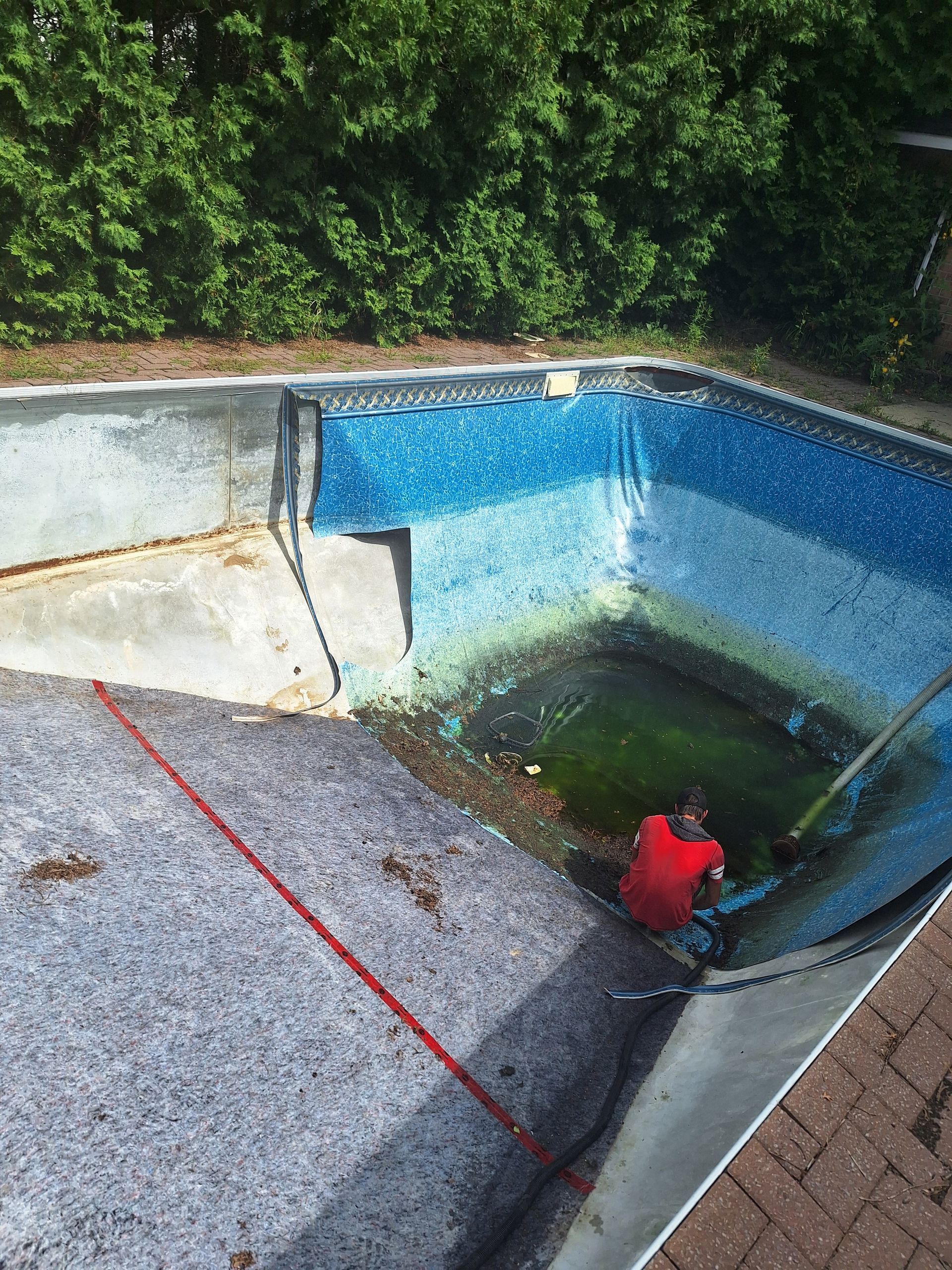 Une personne vêtue d'une chemise rouge travaille dans une piscine vidée et partiellement revêtue, dont l'eau est verte dans la partie profonde.
