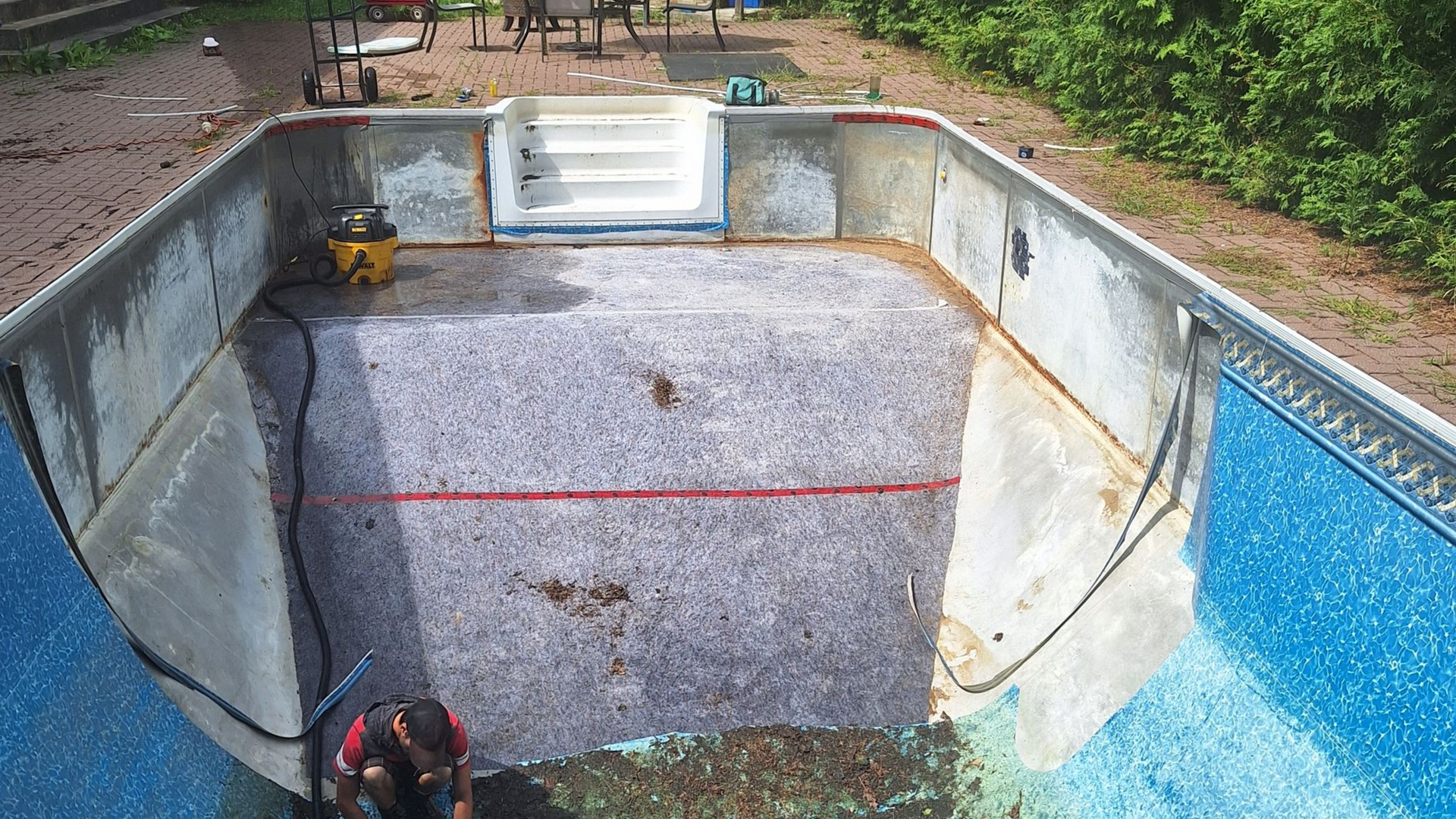 Un ouvrier se trouve à l'intérieur d'une piscine rectangulaire enterrée vide, en cours de rénovation ; on aperçoit les revêtements bleus et les marches blanches.