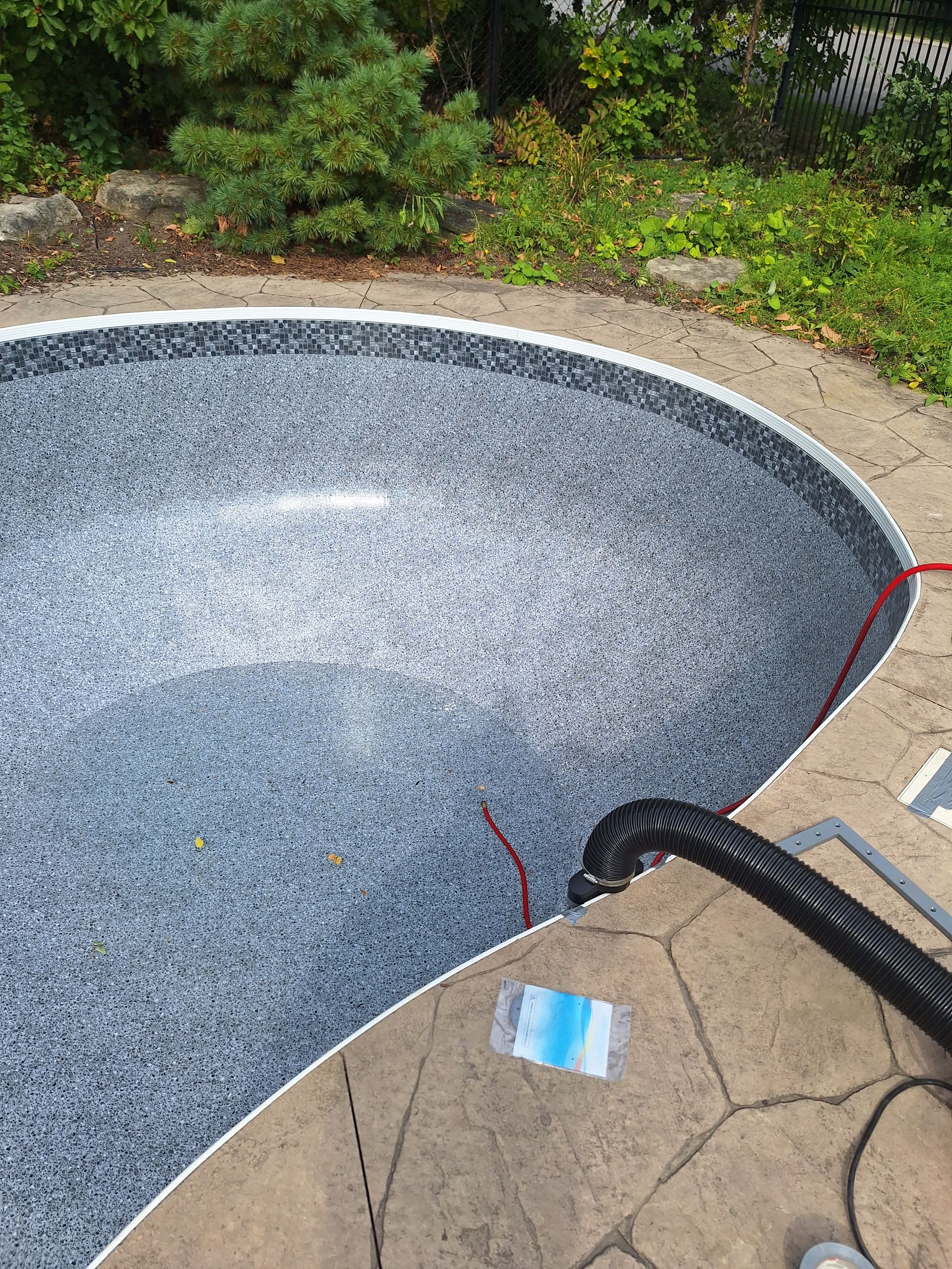 Piscine vide avec revêtement gris moucheté, entourée d'une terrasse en pierre et d'un jardin, avec un tuyau noir et un cordon rouge.