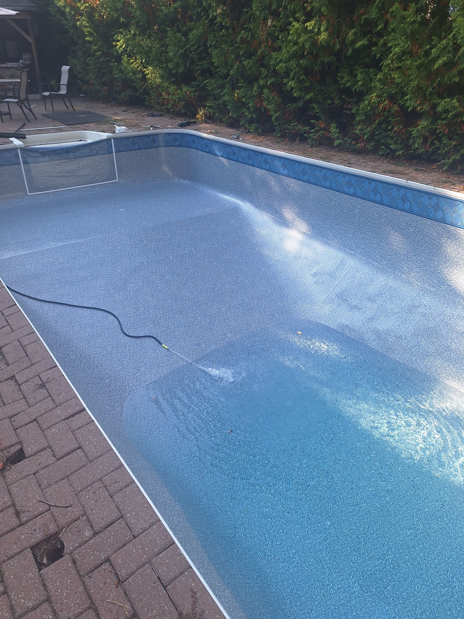 Une piscine rectangulaire vide, revêtue de carreaux bleus à motifs, entourée d'une terrasse en briques et d'une haie feuillue.