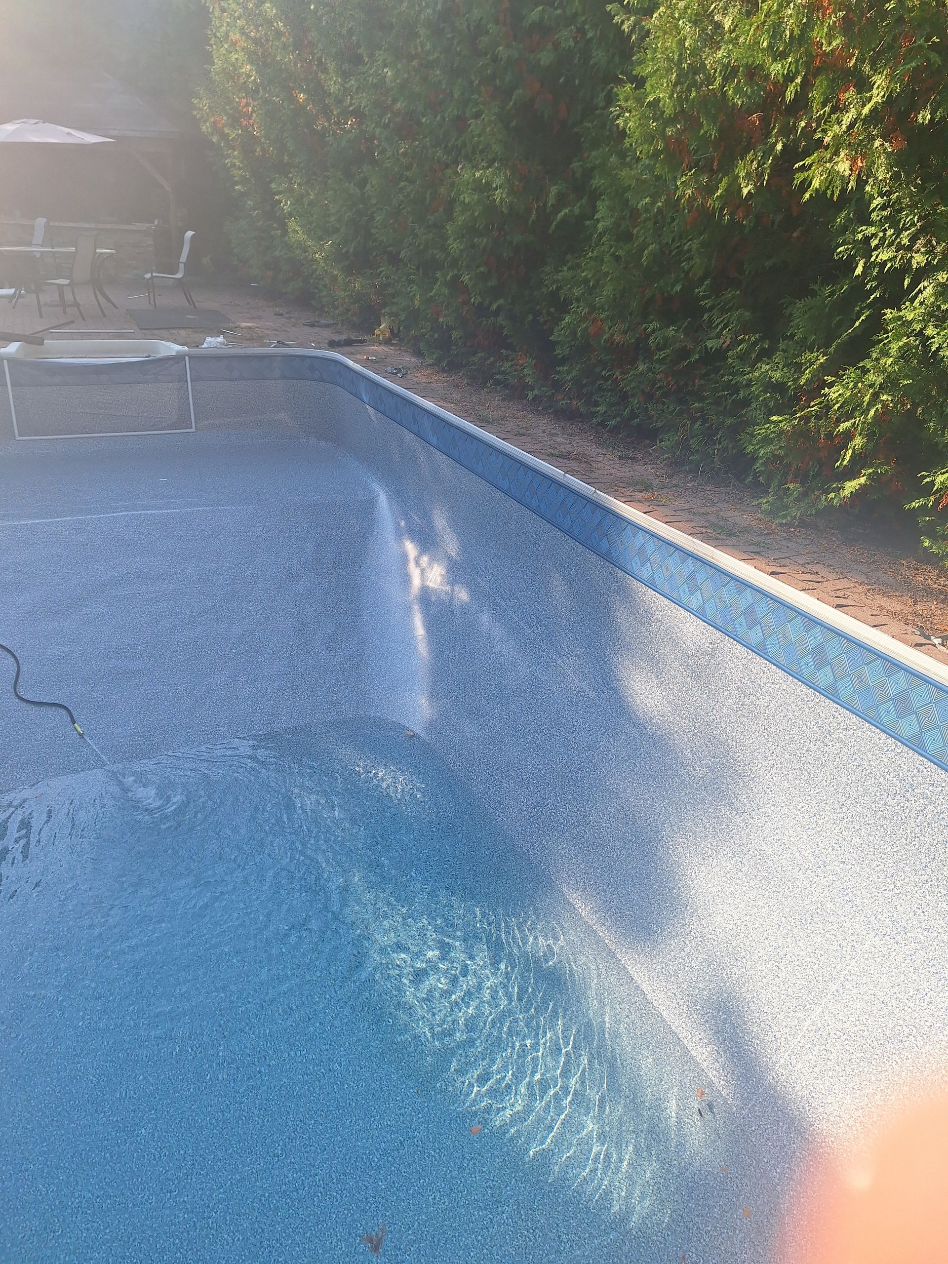 Une piscine rectangulaire vide, avec un liner bleu à motifs, bordée d'un chemin de gravier et de haies vertes.