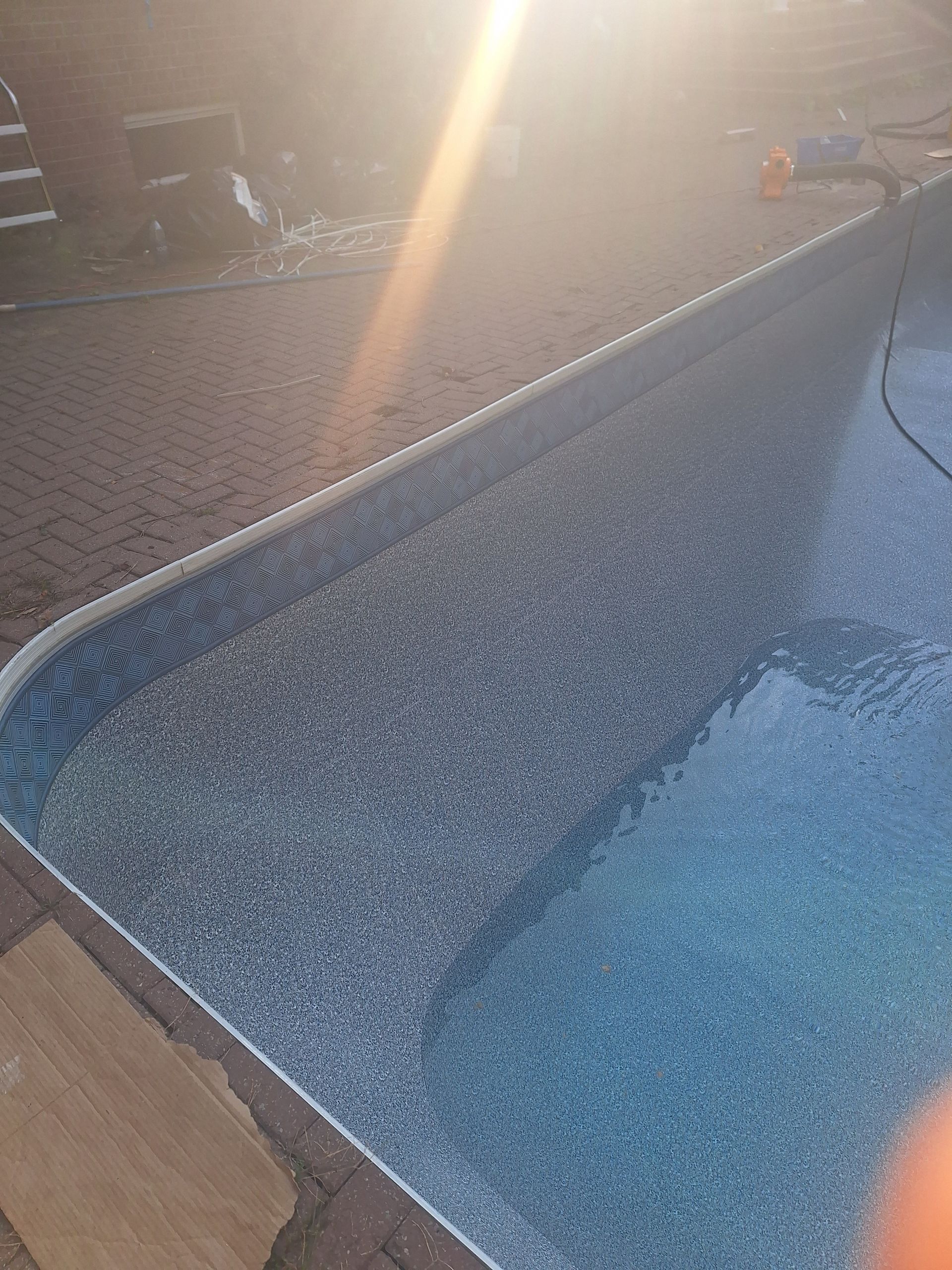 Le bord d'une terrasse de piscine à la finition texturée grise et mouchetée rencontre l'eau bleu clair de la piscine sous un soleil éclatant.