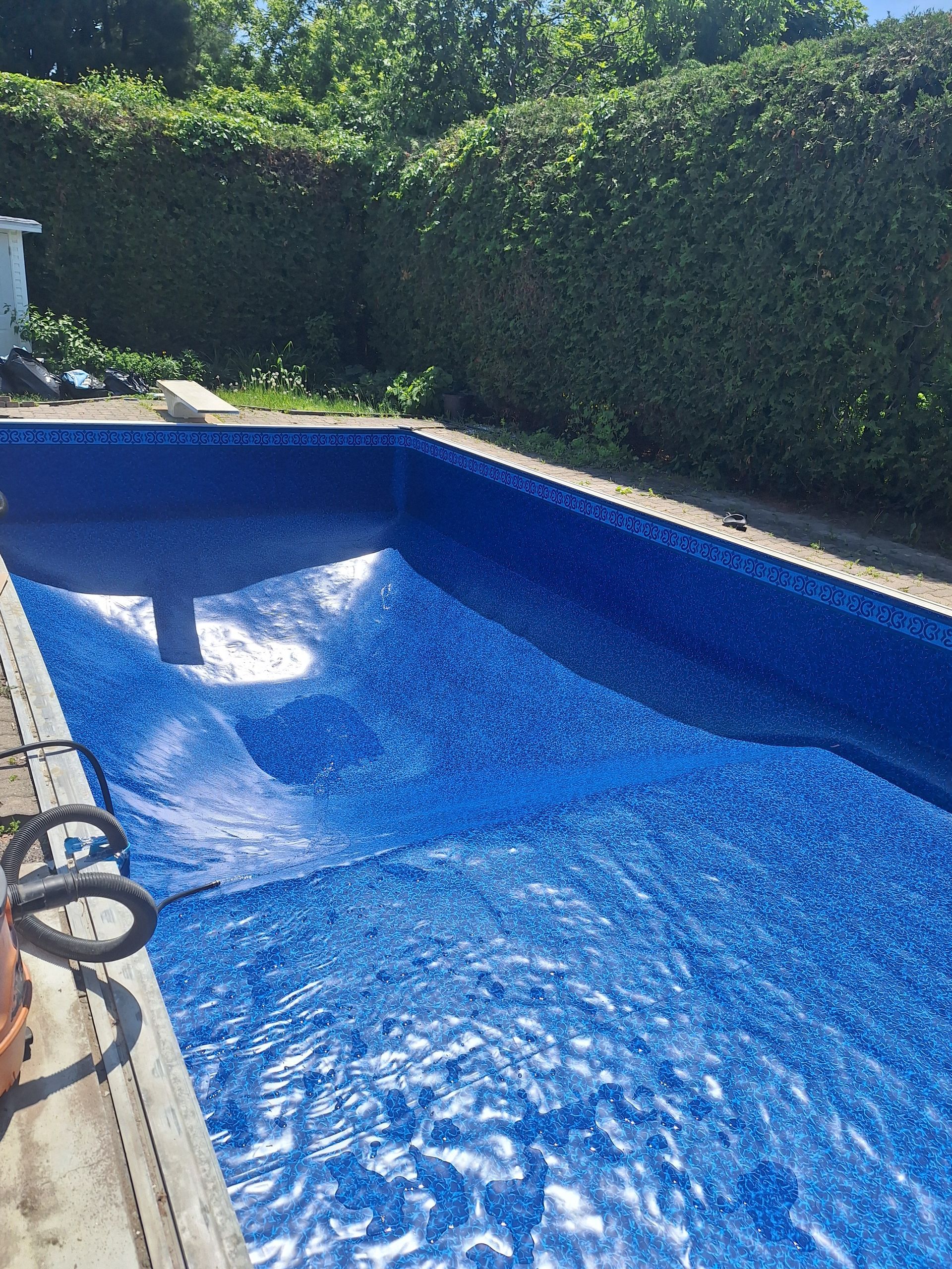 Une piscine rectangulaire à l'intérieur carrelé de bleu, partiellement recouverte d'une bâche bleue, entourée de haies.