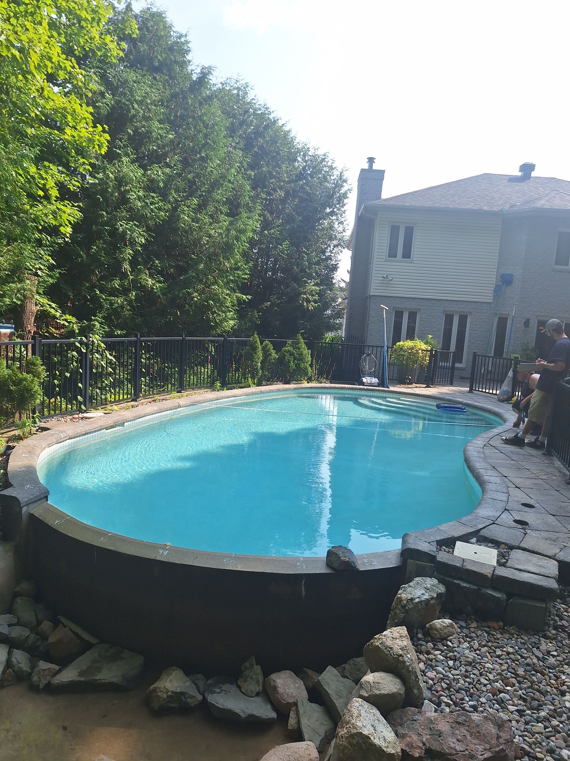 Une piscine ovale hors sol, bordée de pierres, dans un jardin avec une maison et des arbres en arrière-plan.