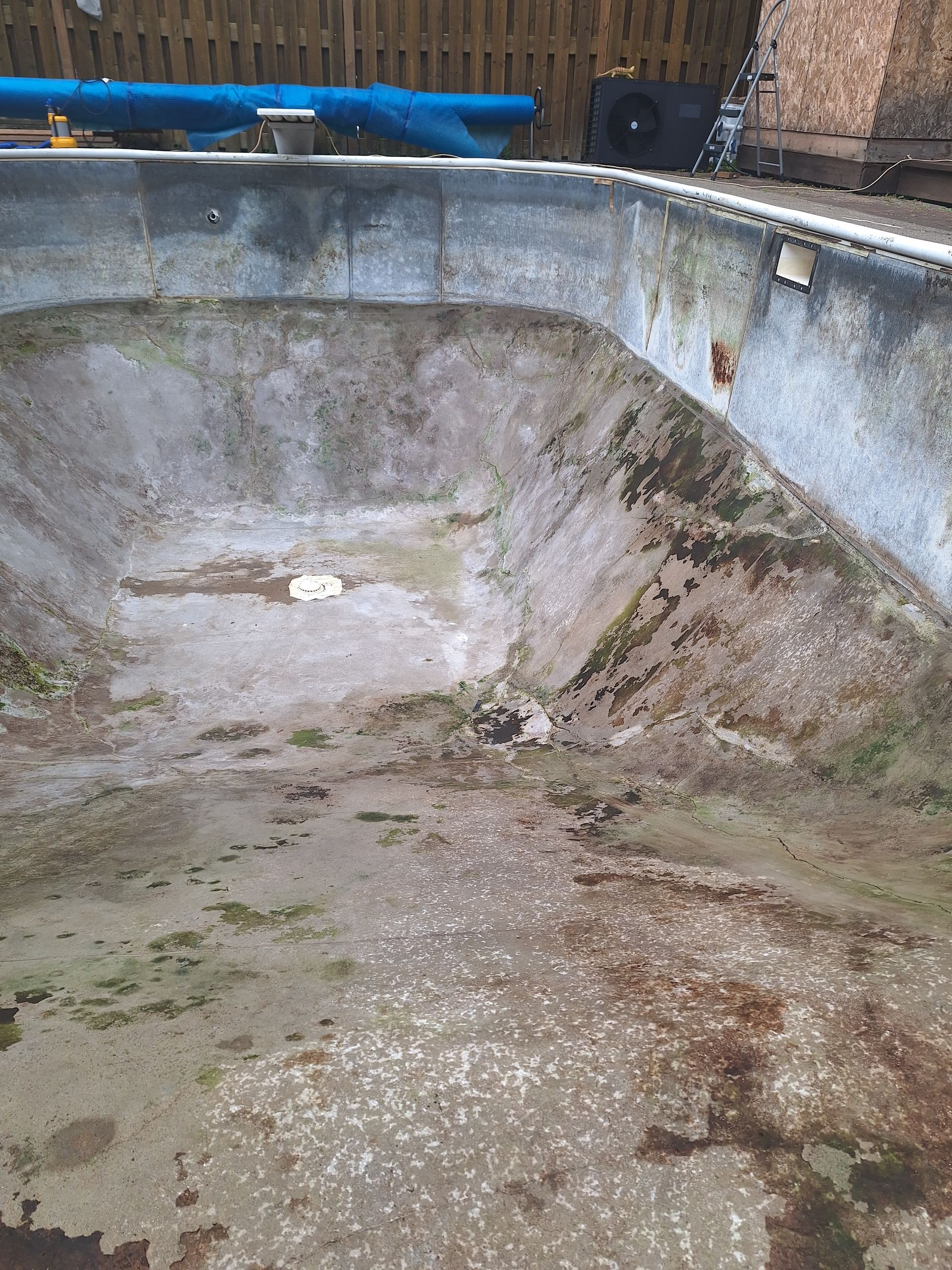 Une piscine en béton vide et délabrée, avec des débris et des taches visibles, se détachant sur un fond de clôture en bois.