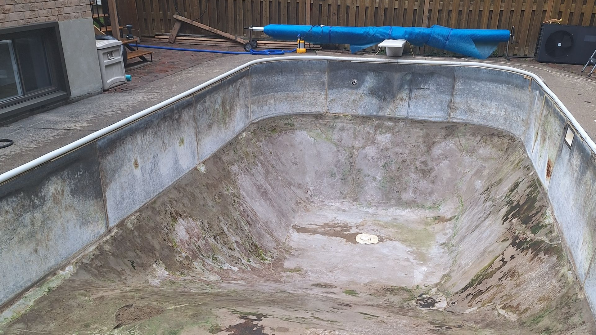 Une piscine enterrée rectangulaire vide, présentant des signes d'usure, située dans un jardin près d'une maison et d'une clôture en bois.