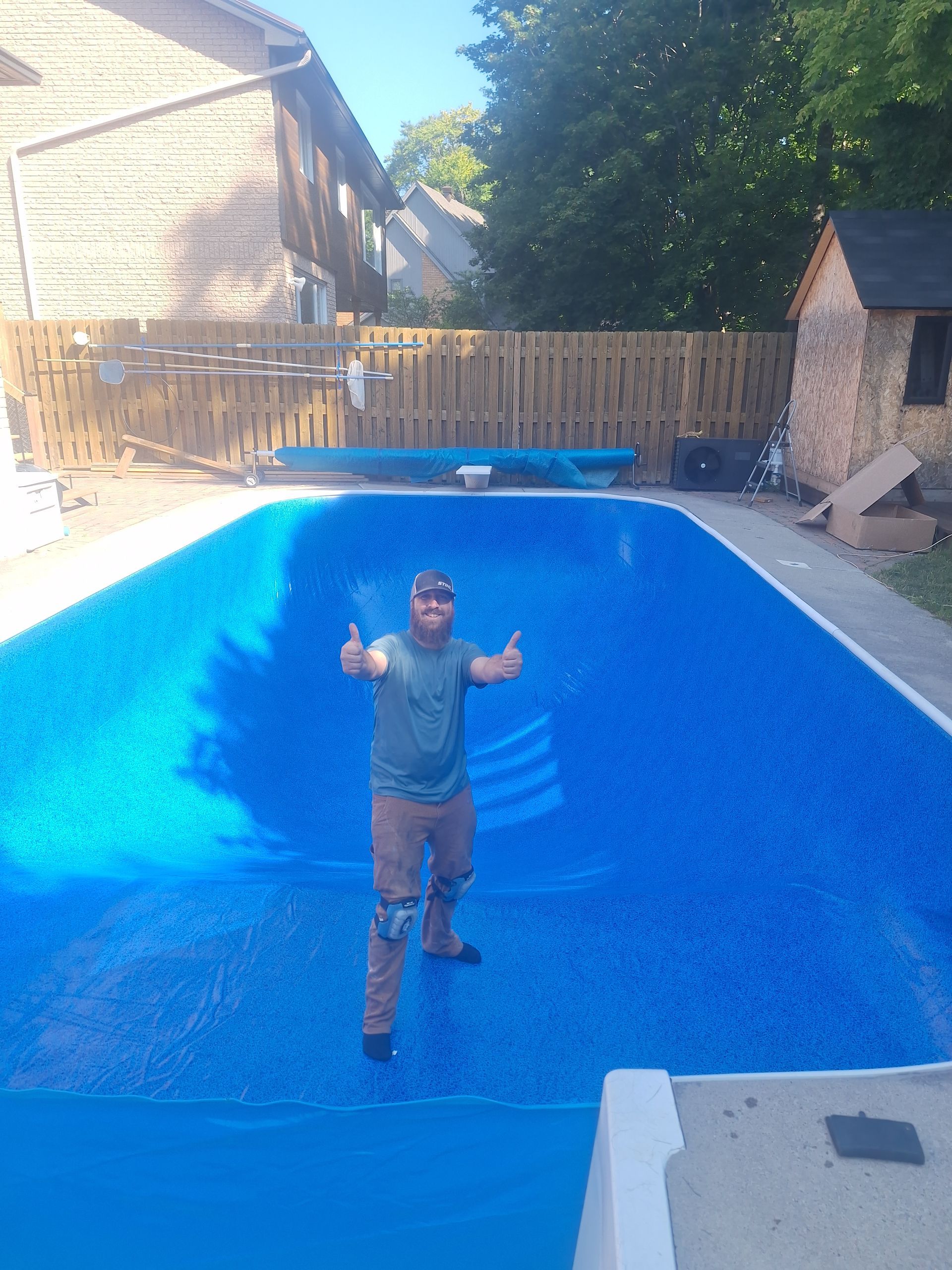 Une personne se tient debout dans une piscine vide d'un bleu éclatant, dans un jardin, et lève les deux pouces tout en portant des vêtements de travail.