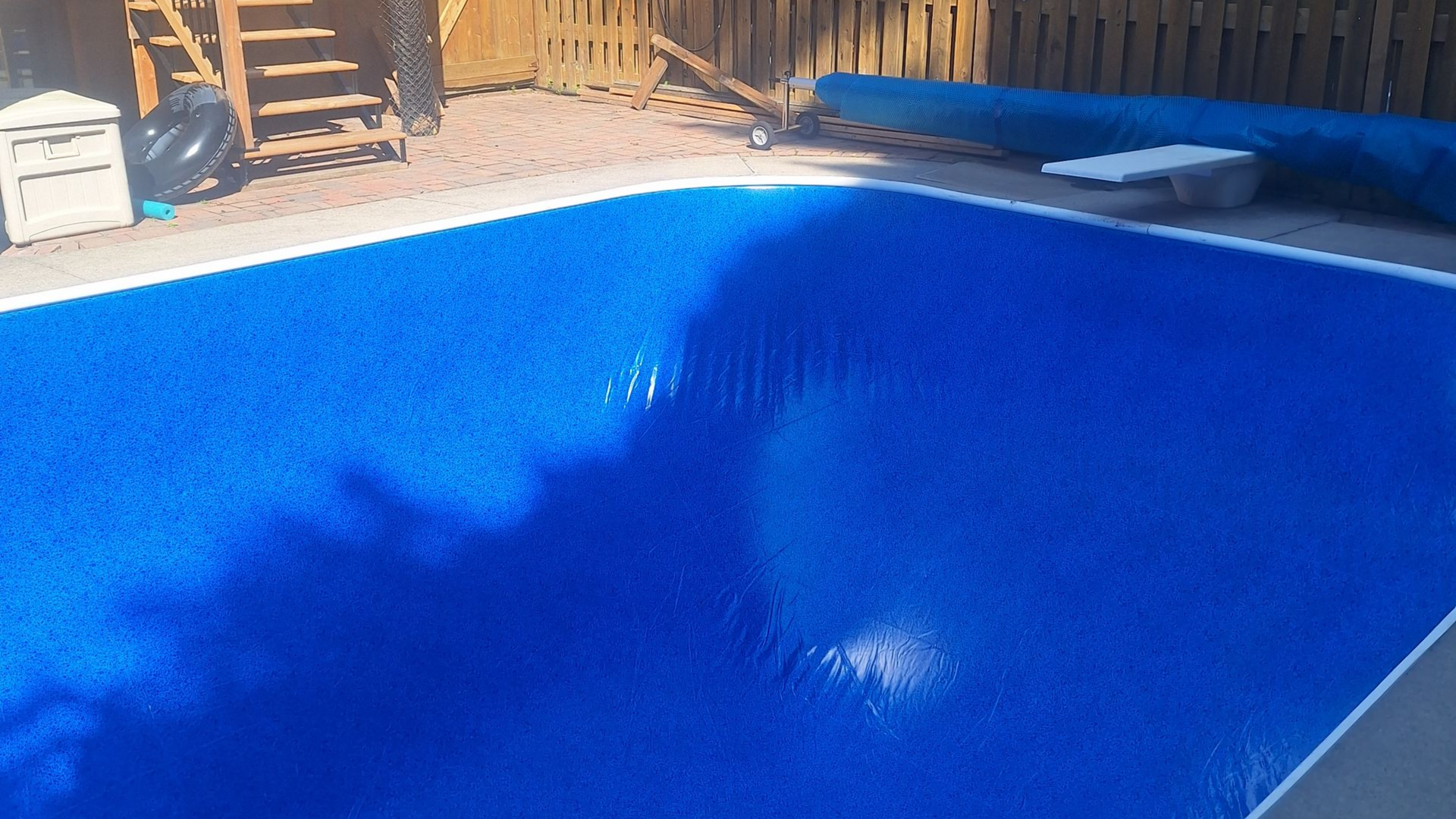 Une piscine rectangulaire avec une couverture bleue, située dans un jardin près d'une clôture en bois et d'un escalier.