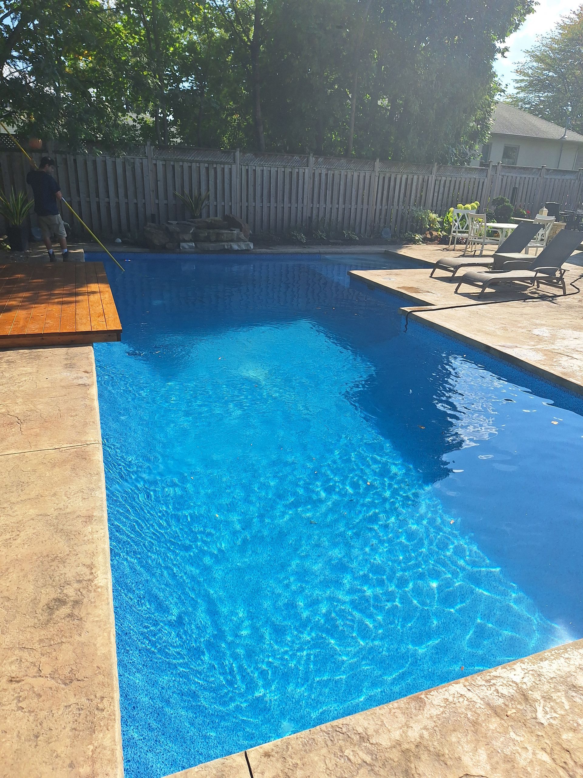 Une piscine rectangulaire bleu vif dans un jardin avec une terrasse en pierre, une marche en bois et une personne utilisant une perche de piscine.