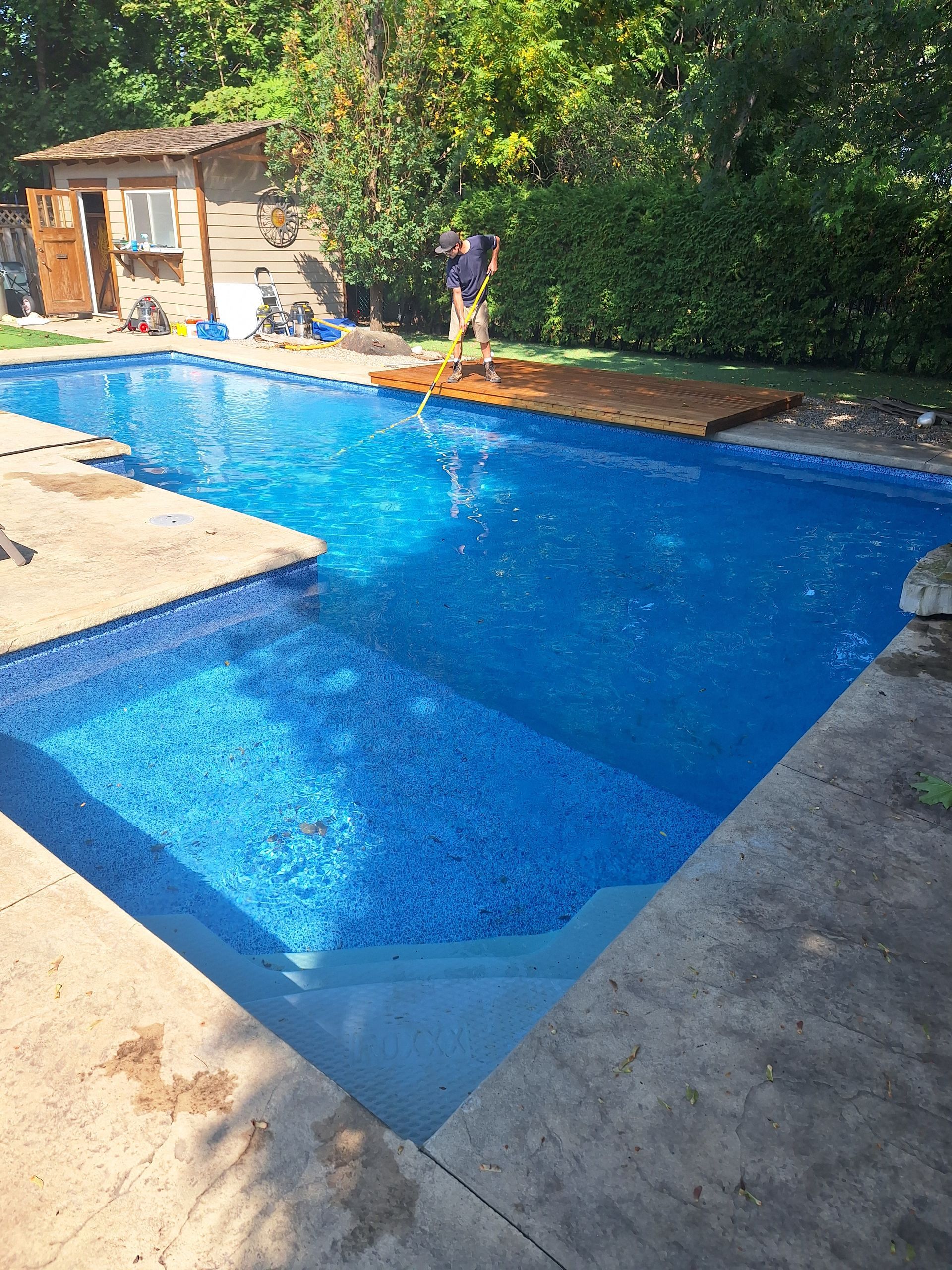 Une personne utilise une perche pour nettoyer une piscine bleue dans un jardin avec un petit abri de jardin et de la verdure.