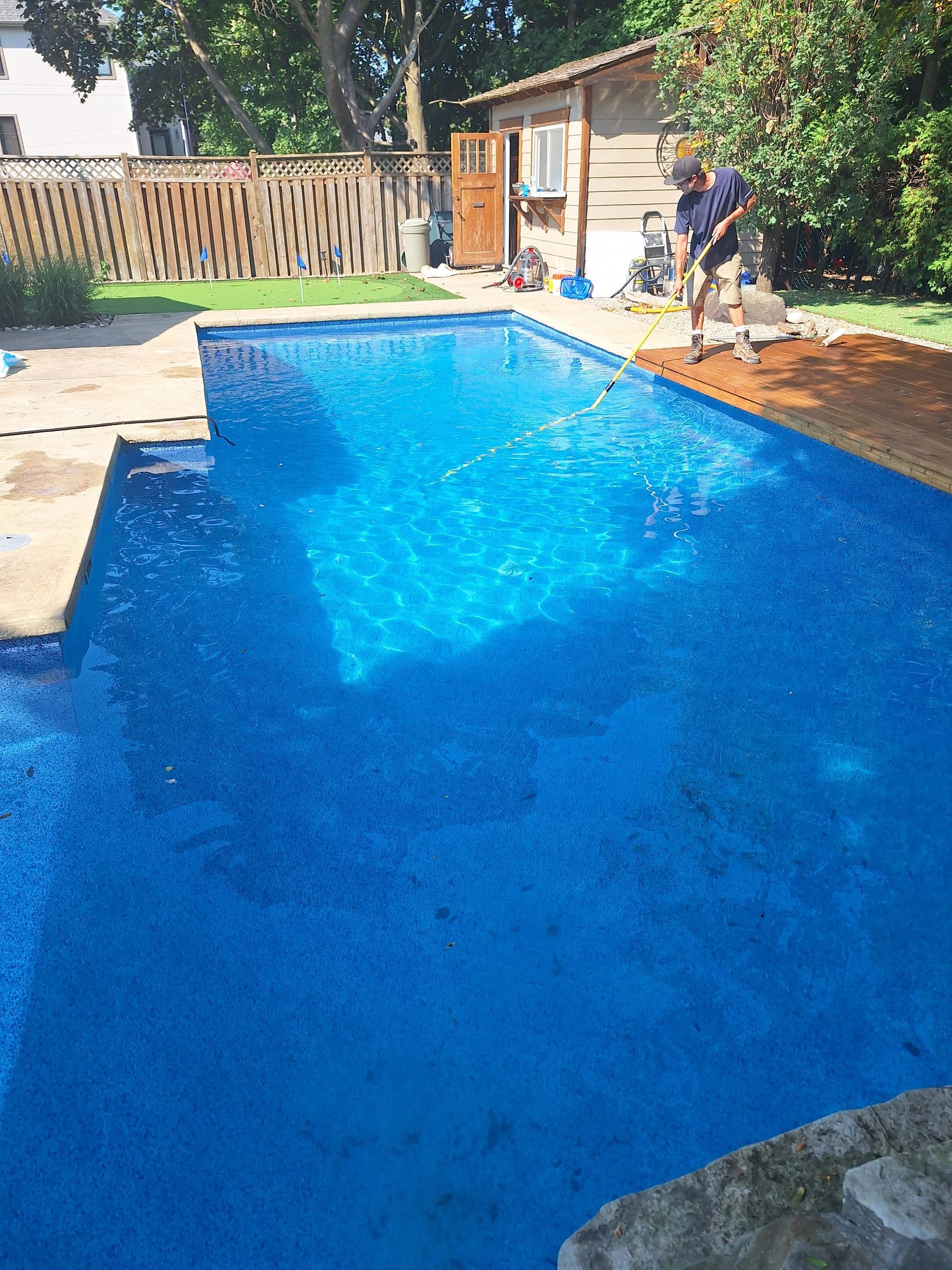 Par une journée ensoleillée, une personne utilise une épuisette à long manche pour nettoyer les feuilles d'une piscine d'un bleu éclatant située dans un jardin.