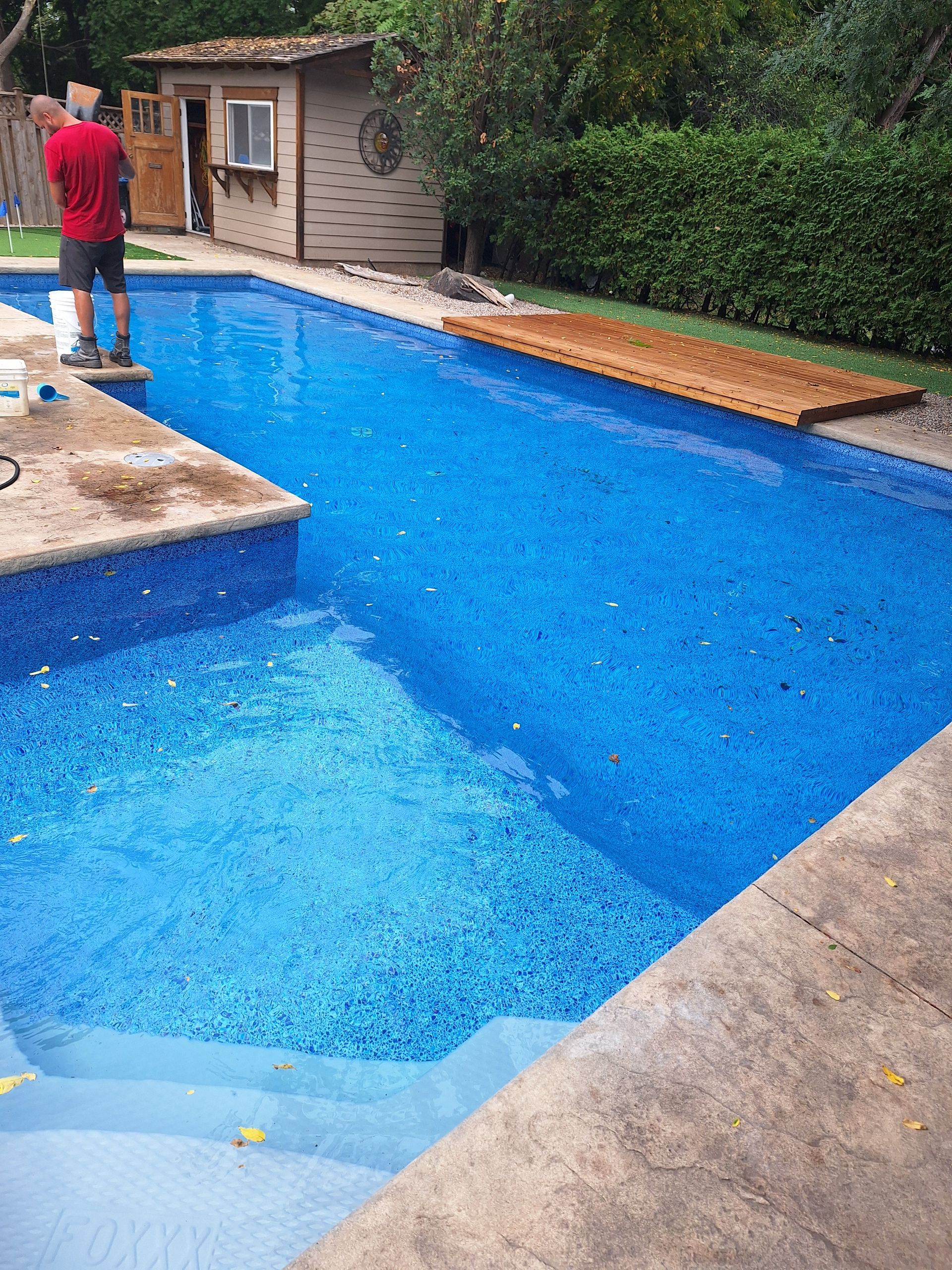 Une personne vêtue d'une chemise rouge se tient au bord d'une piscine bleu vif, avec des escaliers au premier plan et un abri de jardin à proximité.