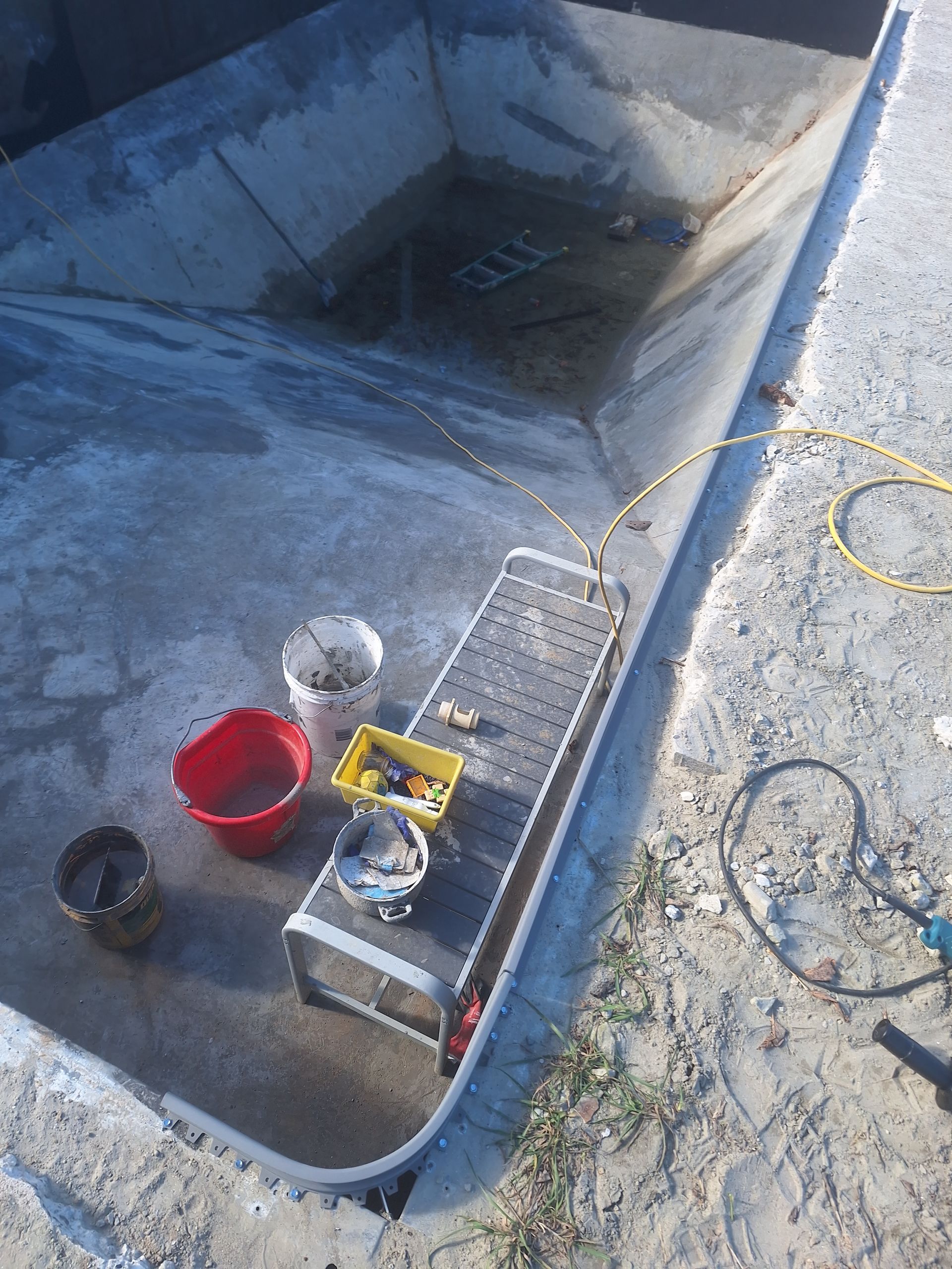 Une piscine en béton en construction, avec un établi métallique sur lequel sont posés des seaux et des outils, et une rallonge électrique à proximité.