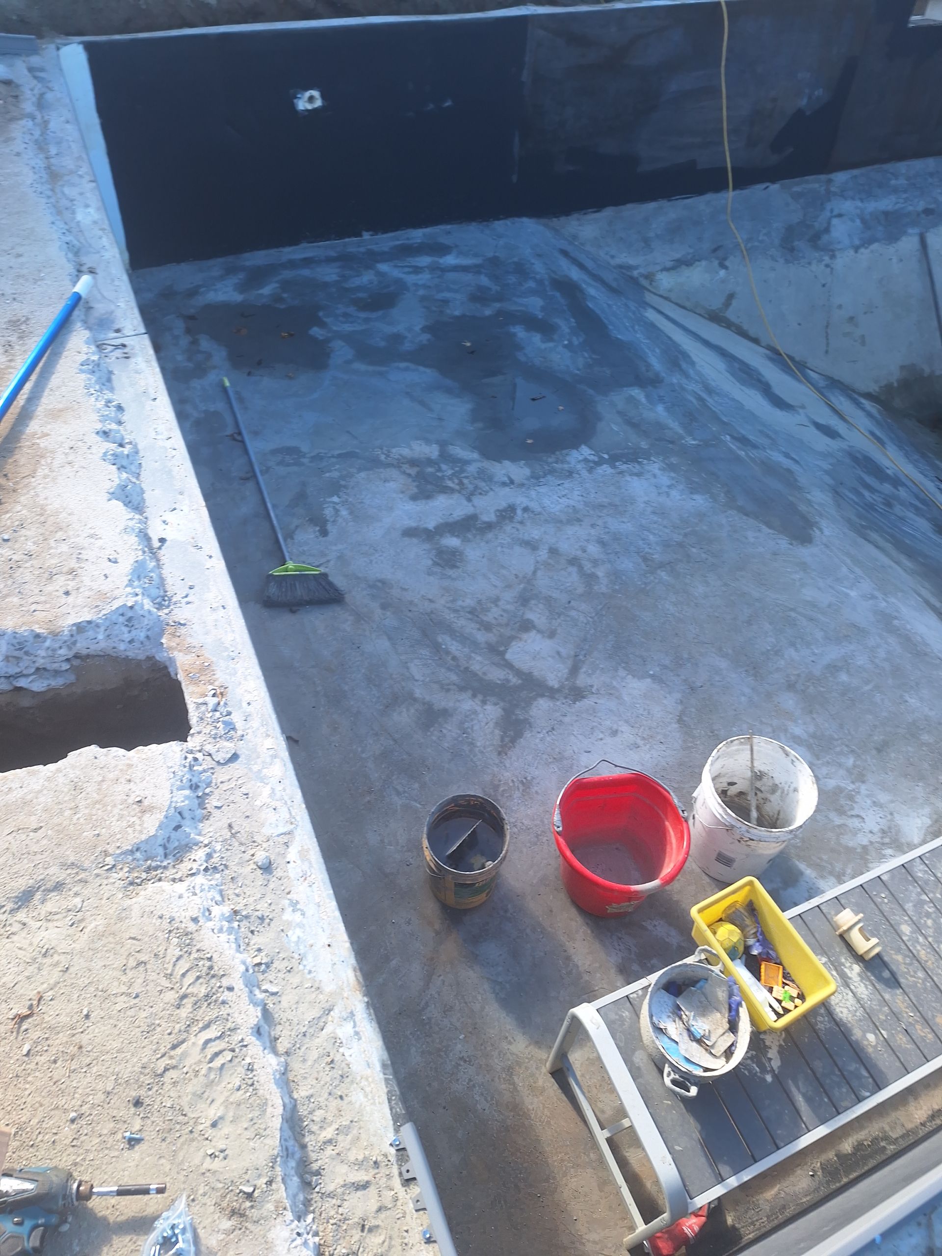 Une piscine partiellement rénovée avec murs et fond en béton, outils, seaux et une plateforme de travail.