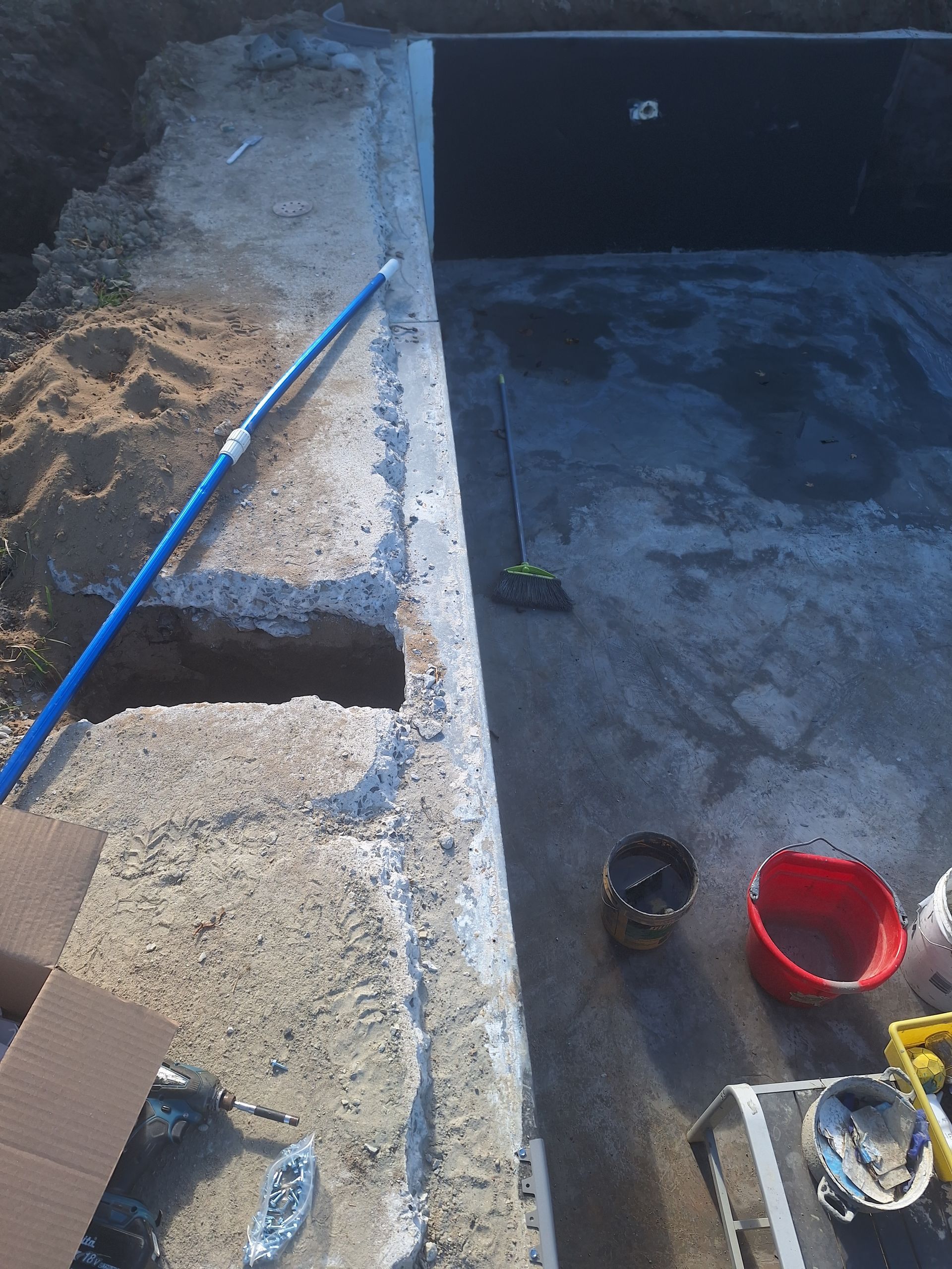 Un chantier de construction de piscine inachevé, avec une corniche en béton, des alentours en terre et du matériel éparpillé.