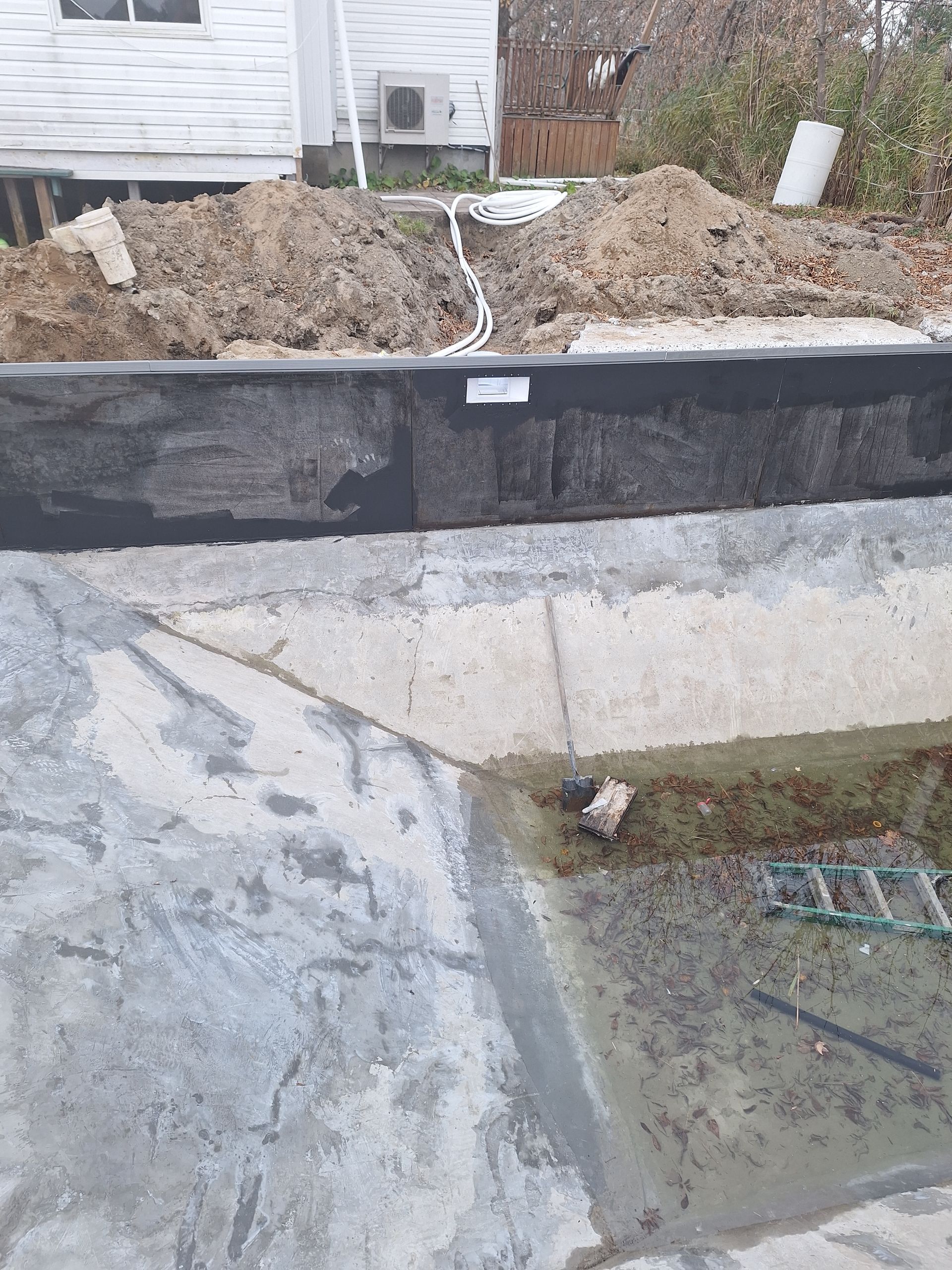 Une piscine en béton en construction avec un mur étanche, de la terre excavée derrière et une maison en arrière-plan.
