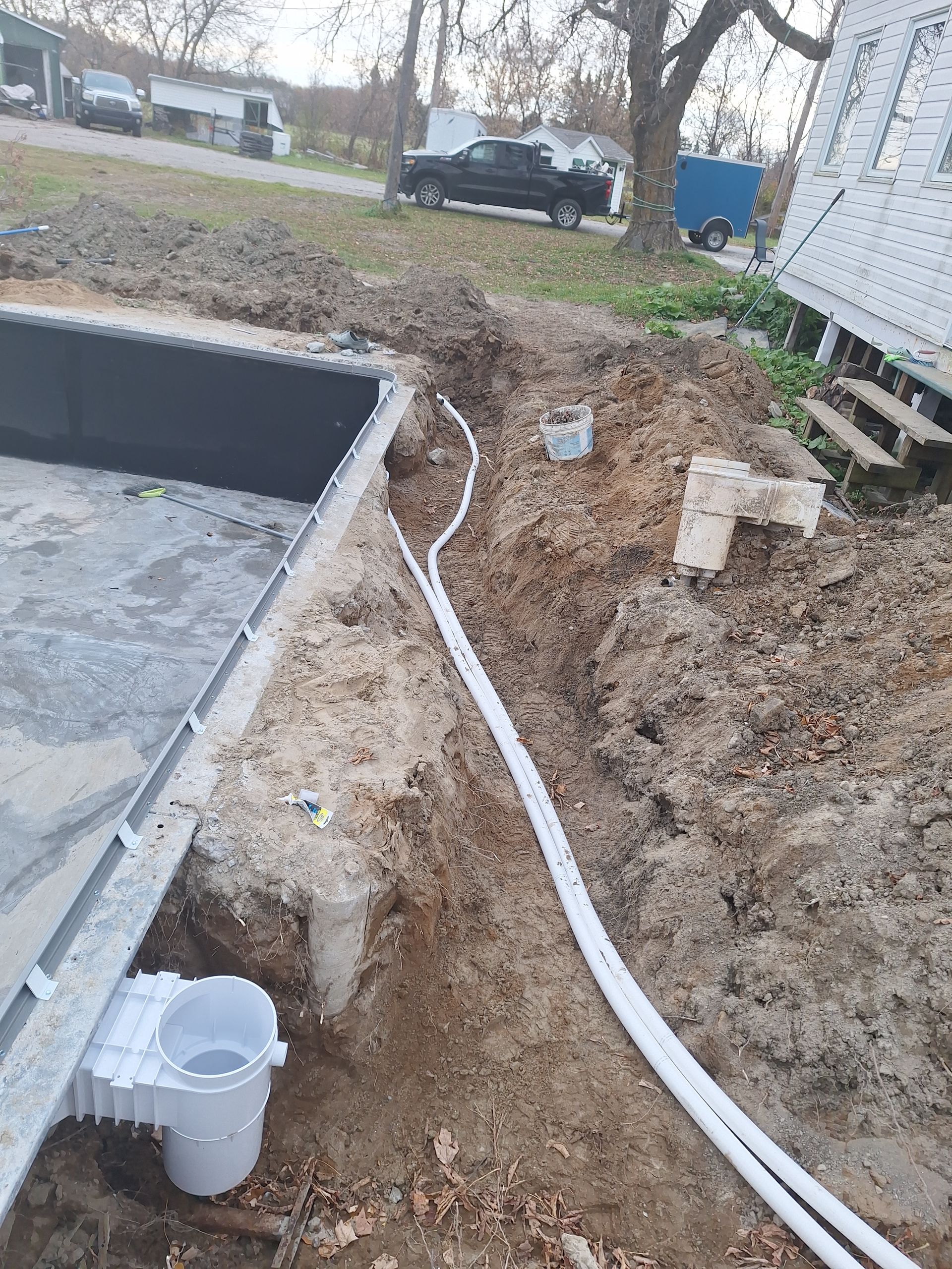Un skimmer de piscine installé sur une paroi sombre, avec des tuyaux blancs traversant une tranchée de terre dans un jardin.