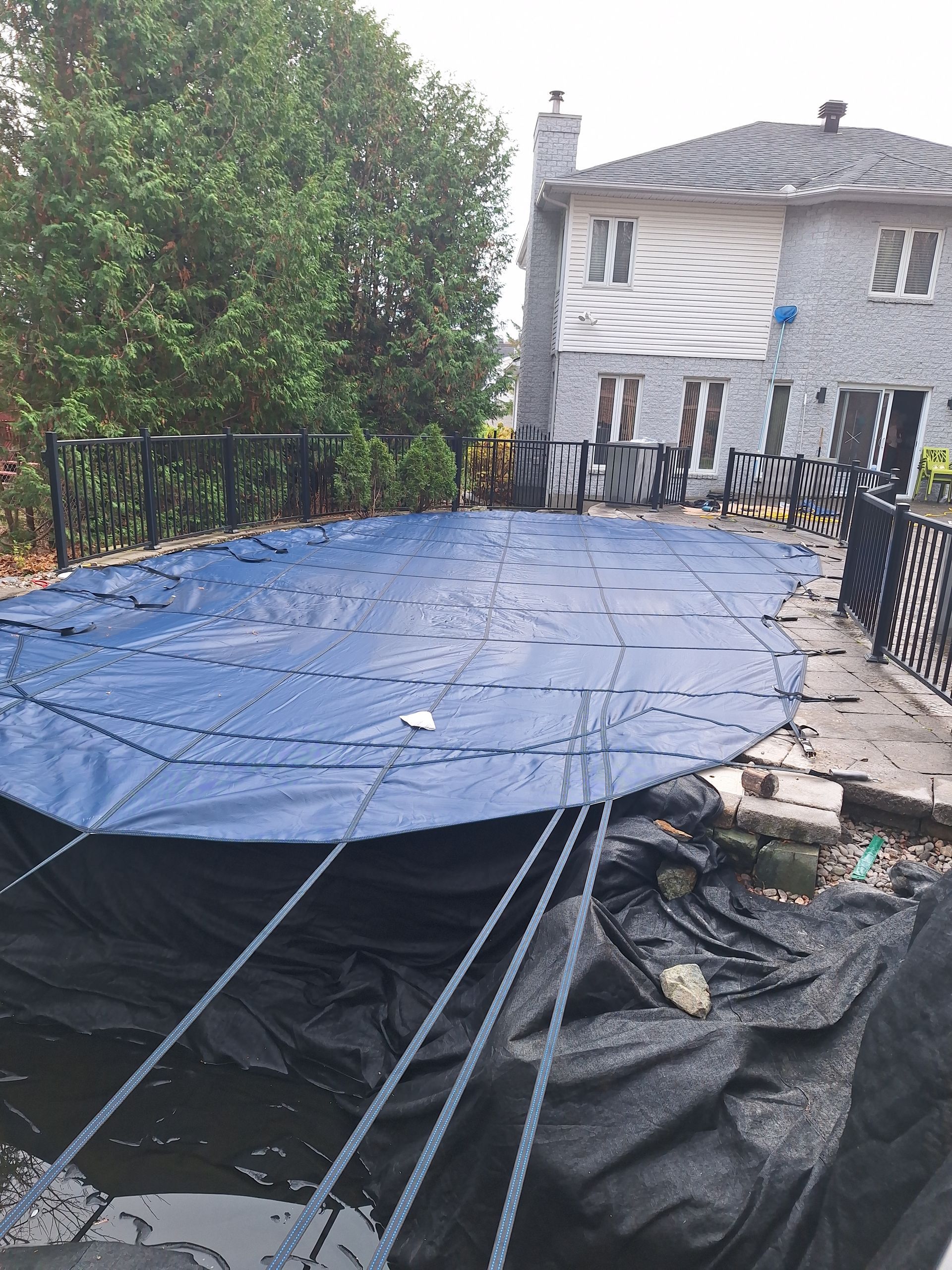 Une piscine ovale recouverte d'une bâche bleue, fixée par des cordes et entourée d'une clôture et d'une maison grise à deux étages.
