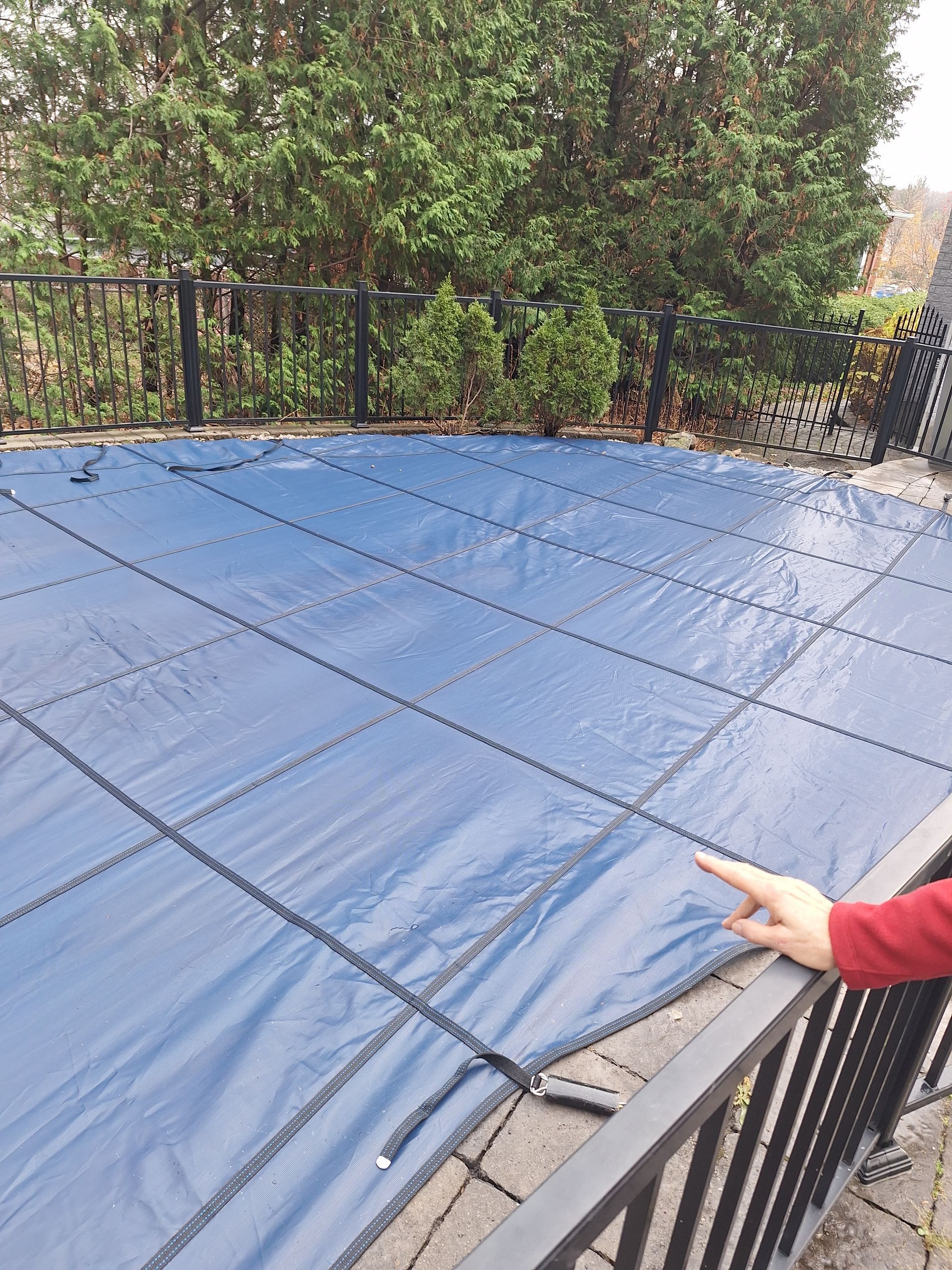 La main d'une personne désigne une bâche de piscine rectangulaire bleue, fixée par des sangles foncées, sur une terrasse près de la végétation extérieure.
