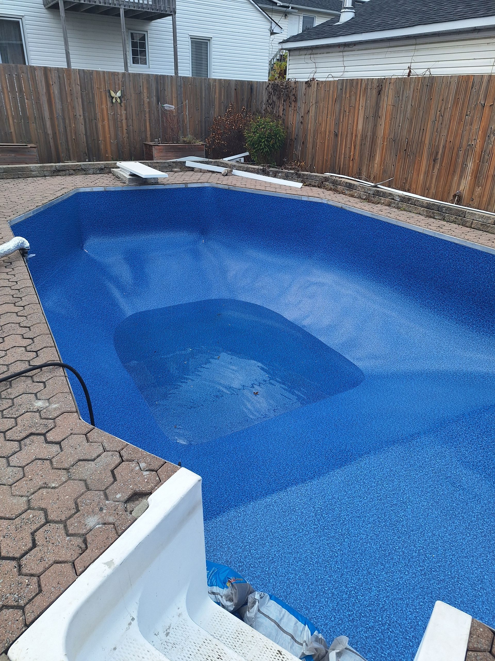 Une piscine vide dans une cour arrière, ornée de carreaux bleu vif et de marches blanches, entourée d'une terrasse pavée et d'une clôture.