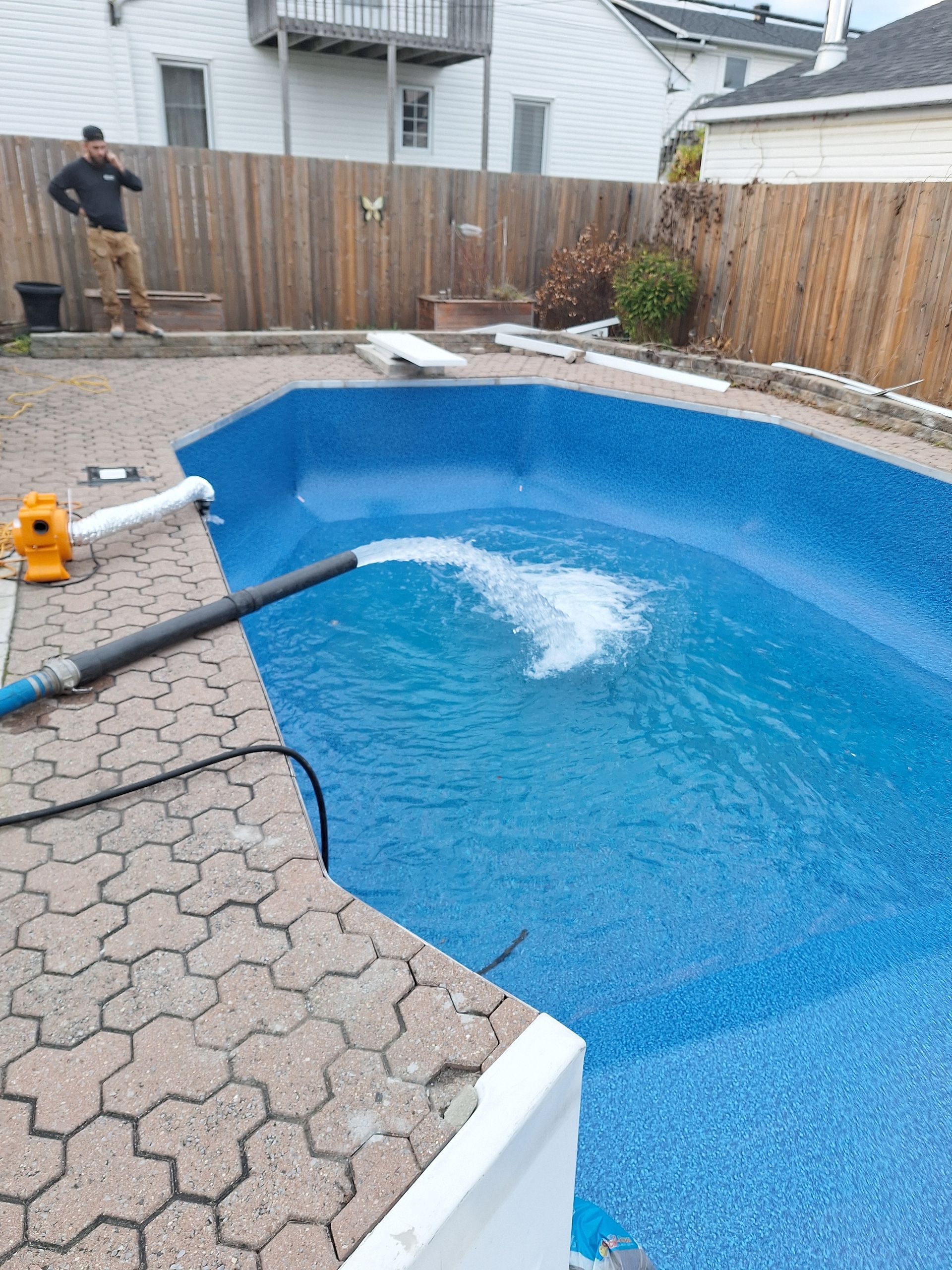 Une piscine aux parois bleues est remplie d'eau à l'aide d'un tuyau d'arrosage, une personne se tenant sur la terrasse en arrière-plan.