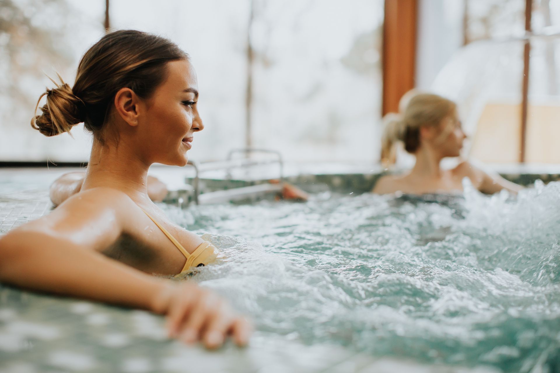 Two people relaxing in a bubbling hot tub in a sunlit, indoor spa setting.