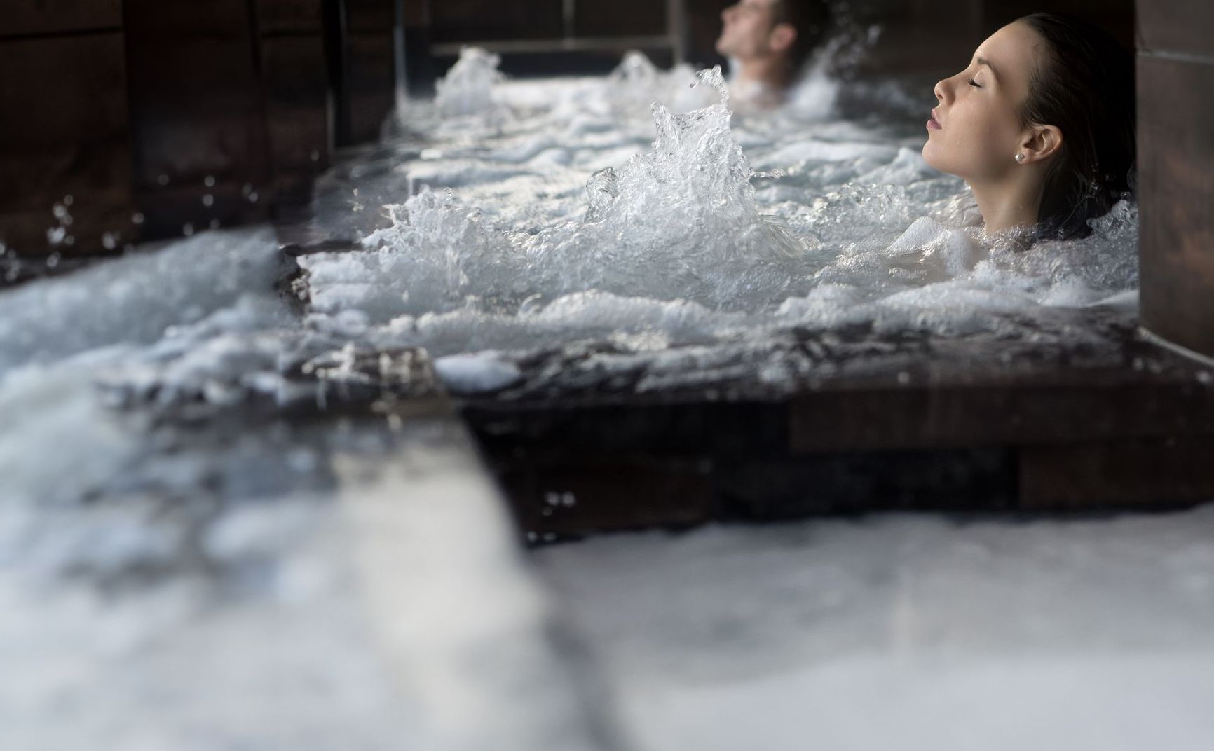 A person relaxing in a bubbling hot tub with water splashing around them in a dimly lit, tiled indoor spa.