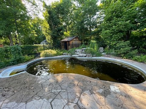 Un étang en forme de rein aux eaux troubles, entouré d'une terrasse en pierre, d'arbres verdoyants et d'un petit abri en bois.