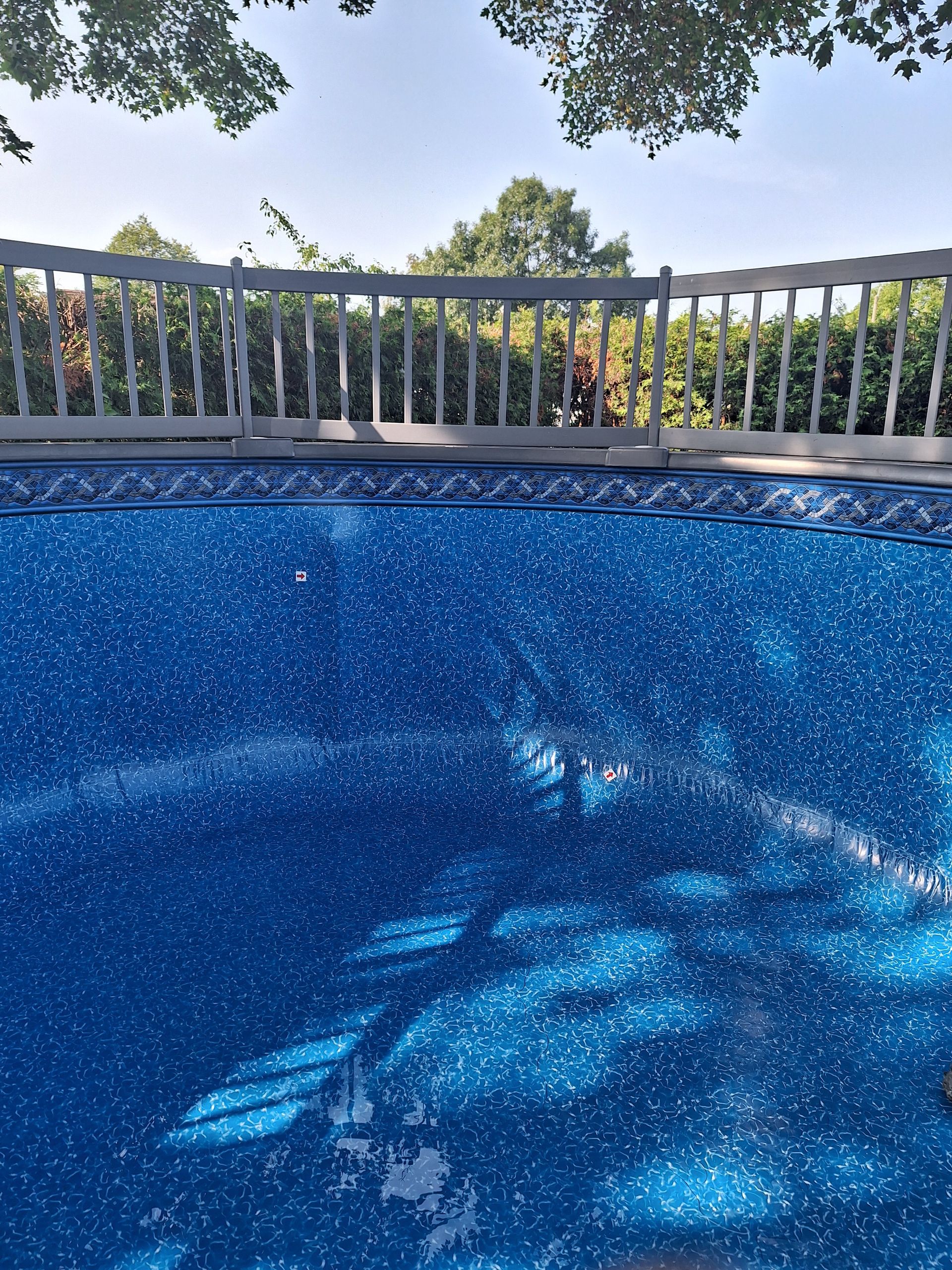 A bright blue, speckled vinyl swimming pool liner under a gray deck fence and tree branches.