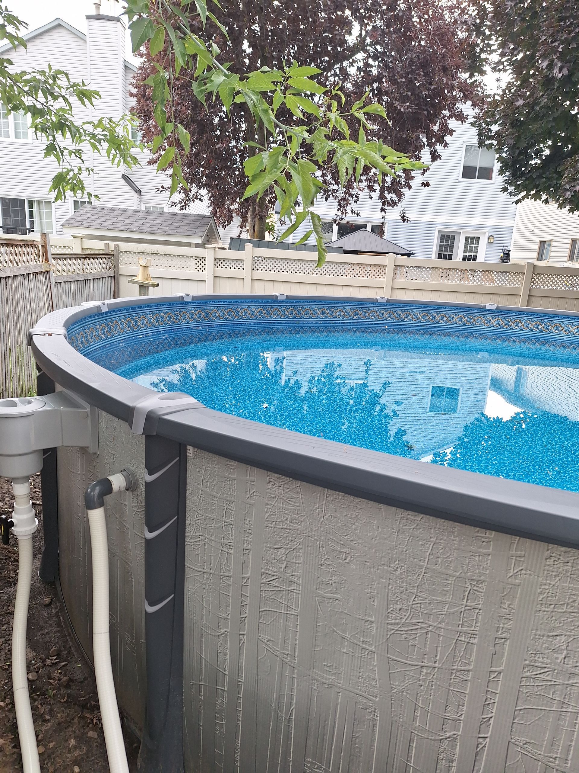Une piscine hors sol ovale, aux parois grises à motifs, avec un rebord supérieur foncé et des tuyaux d'arrivée d'eau, dans un jardin.