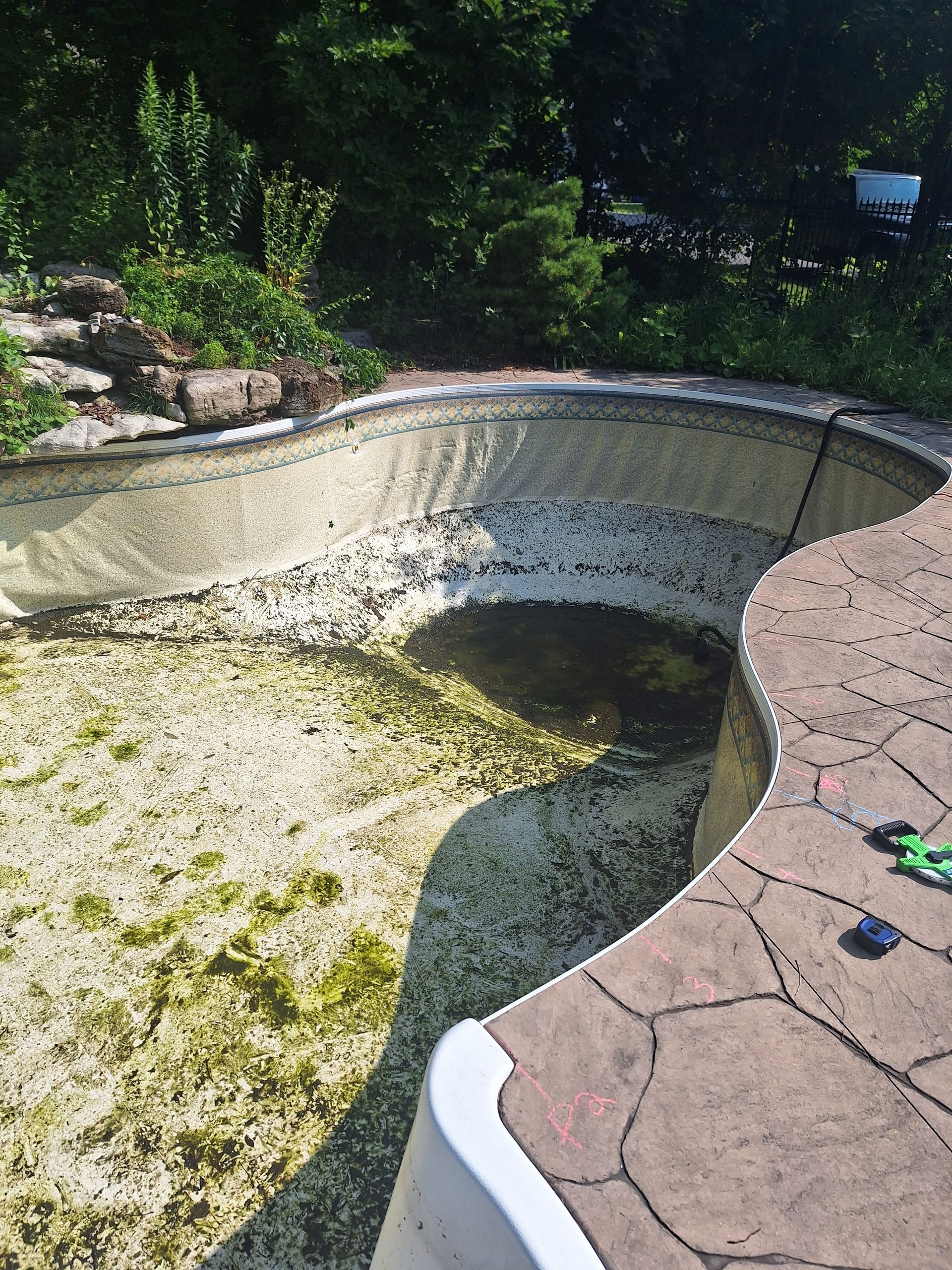 Une piscine vide, remplie de débris, avec une terrasse en pierre dans un jardin.