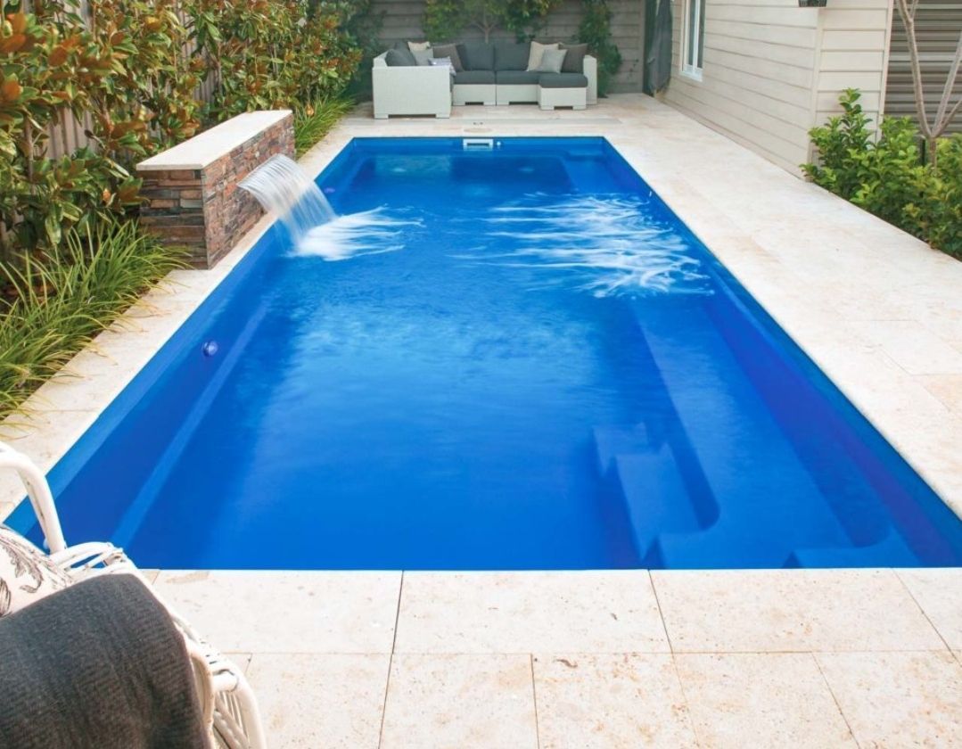 Une piscine rectangulaire bleue avec un bassin en pierre, des sièges extérieurs et une terrasse carrelée de couleur claire.