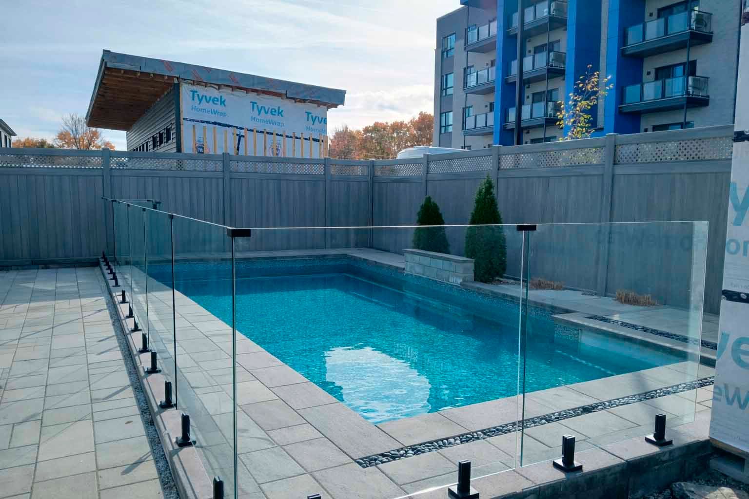 Une piscine rectangulaire à l'eau bleue, dotée d'une barrière de sécurité en verre, située dans un jardin clôturé près d'un bâtiment.