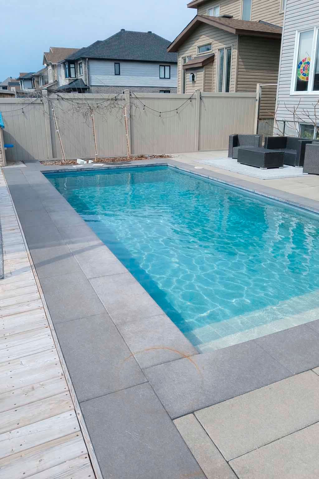 Une piscine rectangulaire dans le jardin, avec une eau bleue et des margelles en pierre grise, entourée d'une terrasse en bois et d'un patio.