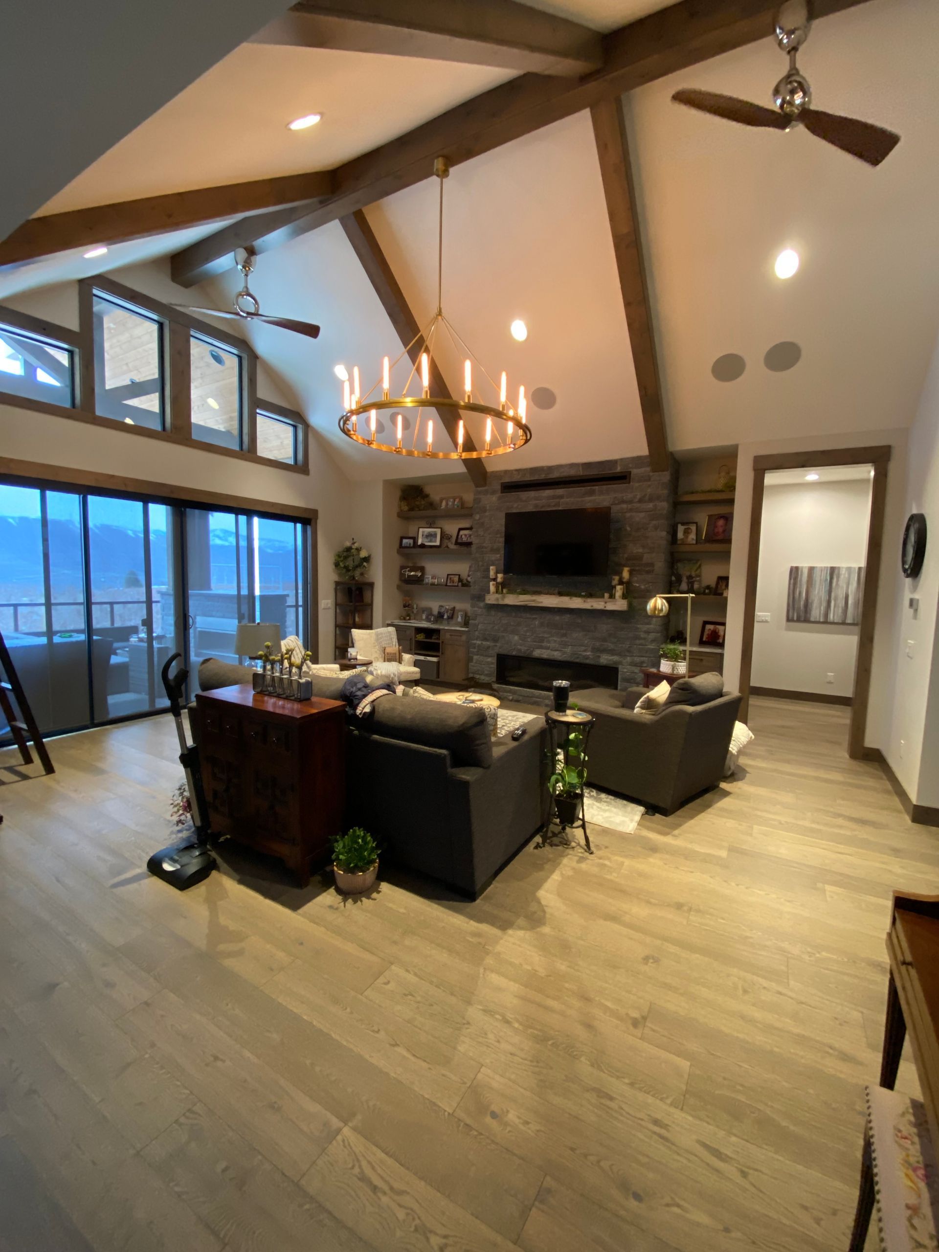 Open-concept living room with stone fireplace, chandelier, ceiling fan, and large windows overlooking a balcony