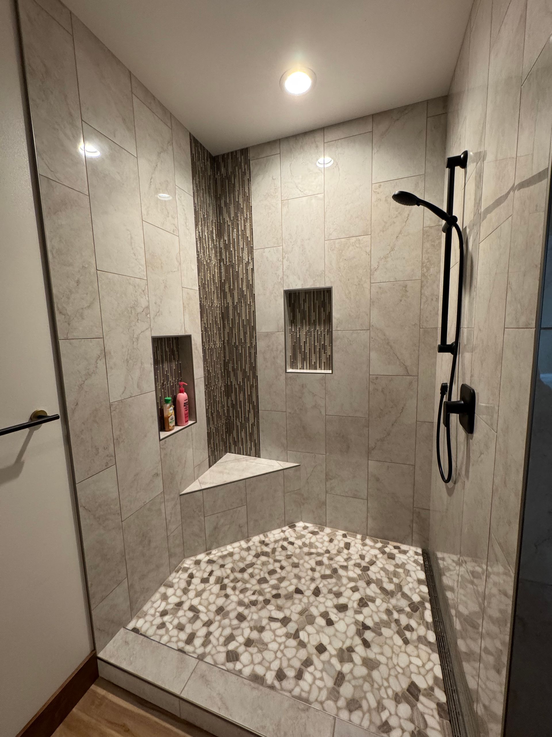 Modern walk-in shower with gray tile, pebble floor, bench, and rainfall showerhead