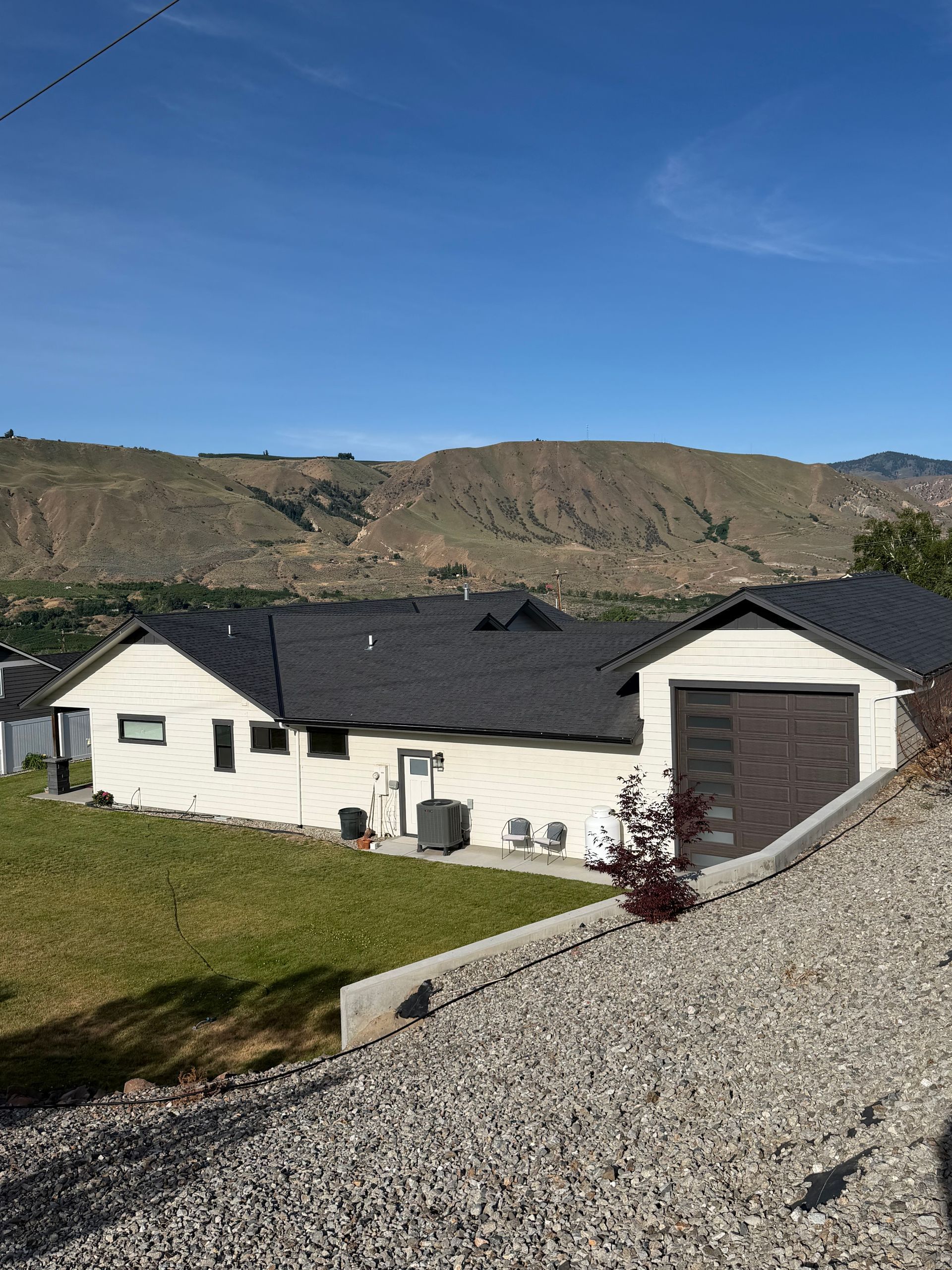 White house with dark roof, detached garage, and gravel driveway in a grassy valley under a clear blue sky