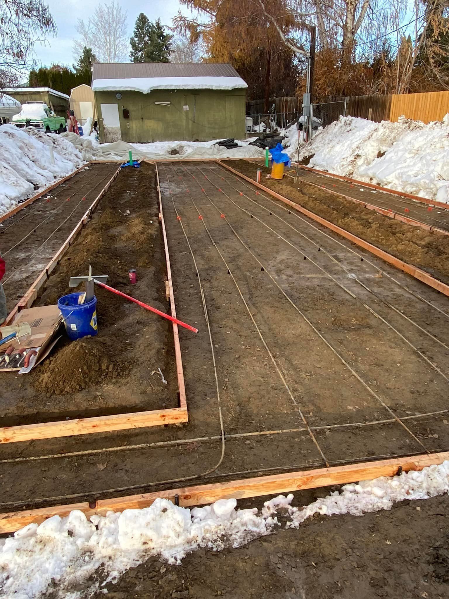Construction site with wooden forms and strings outlining a rectangular area. Dirt, snow, and a green building are visible.