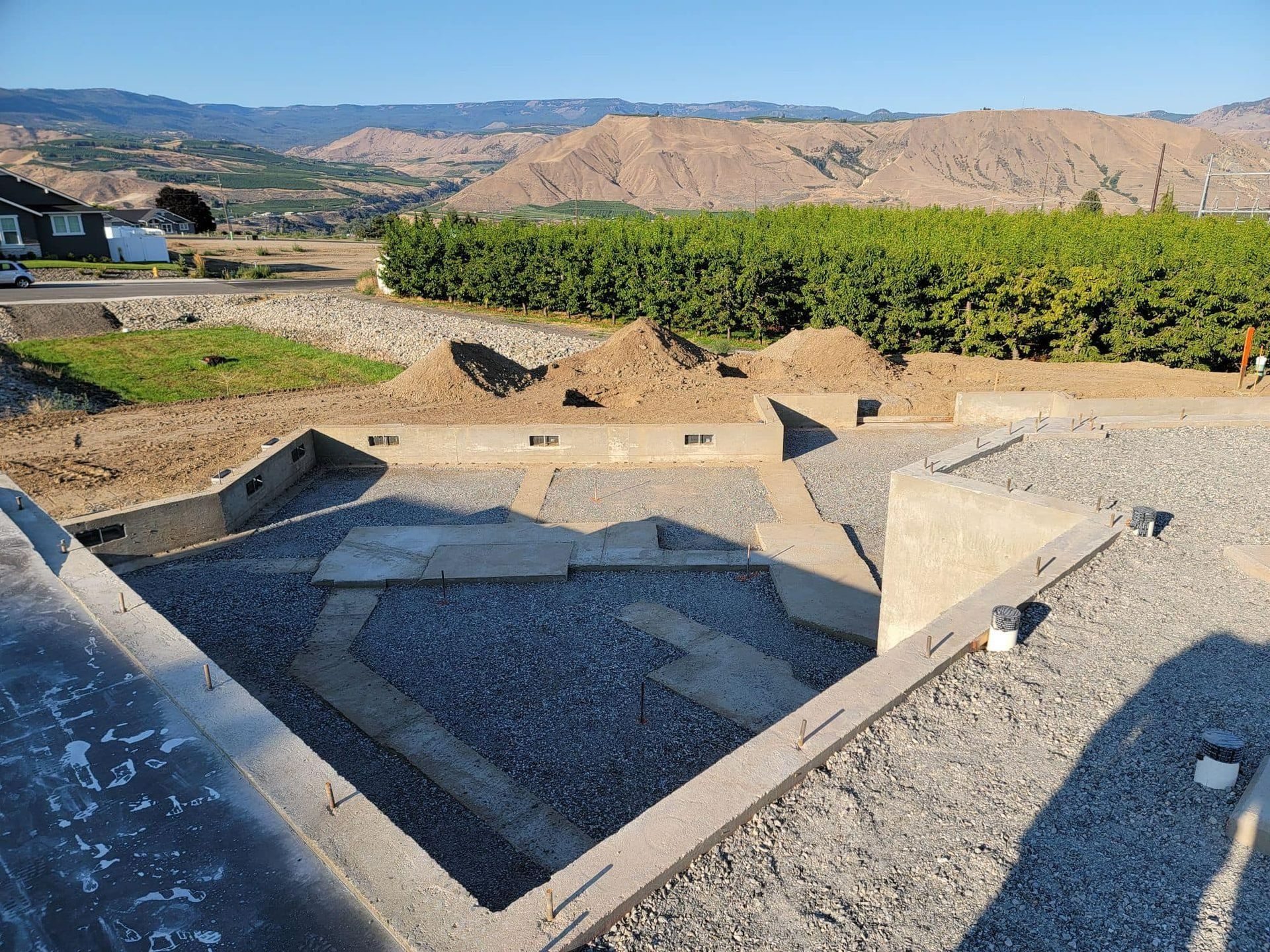 Construction site with concrete foundation and gravel, mountains and vineyard in background.