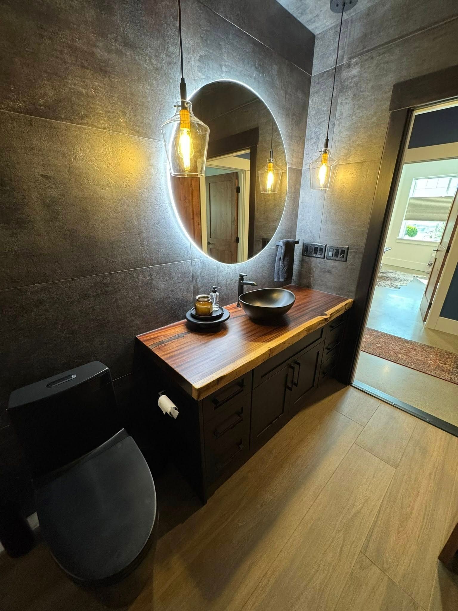 Bathroom with a round, lighted mirror over a dark wood vanity and black toilet.