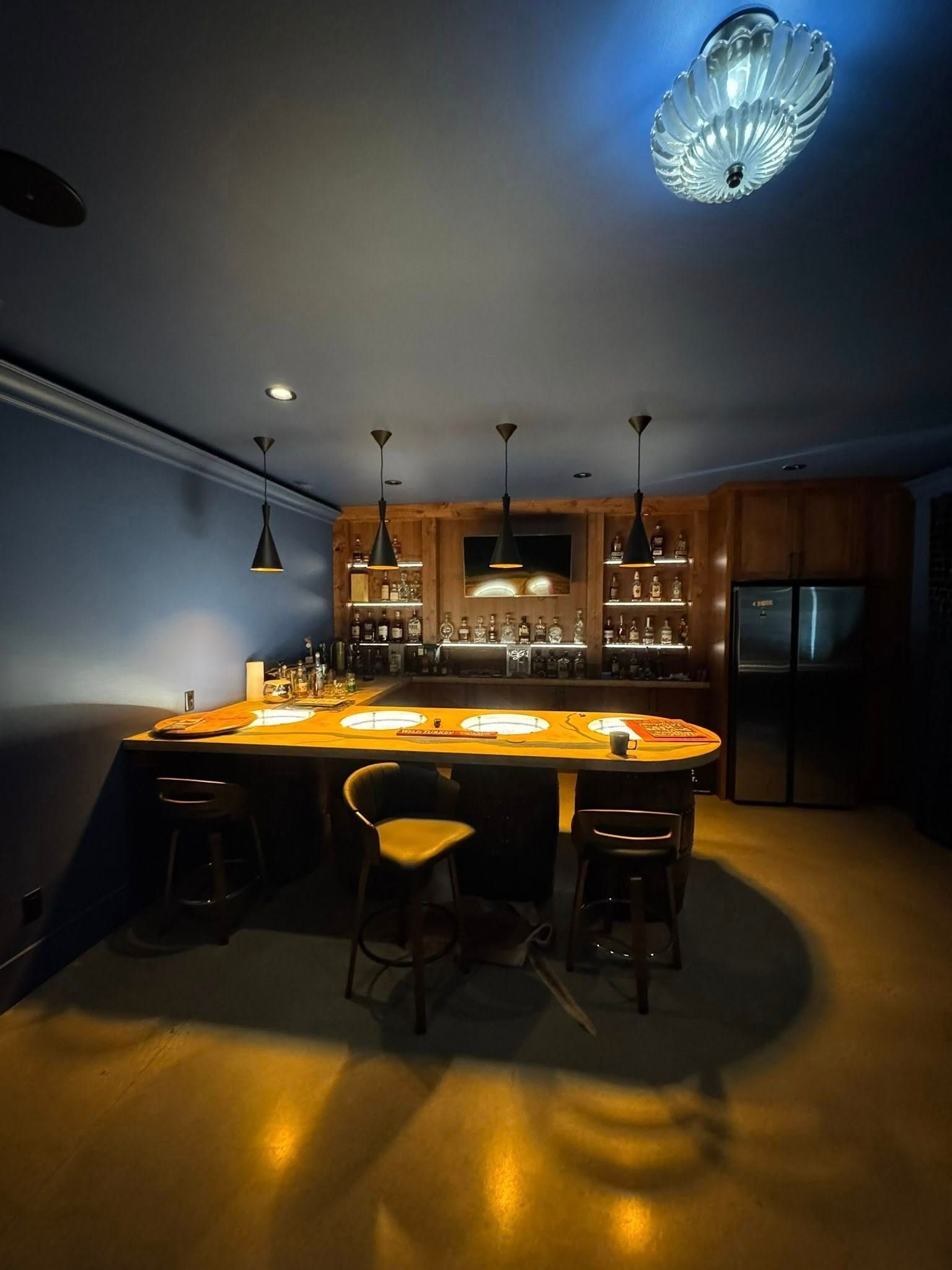 Home bar with wooden counter, stools, shelving, and refrigerator. Soft lighting against blue walls.