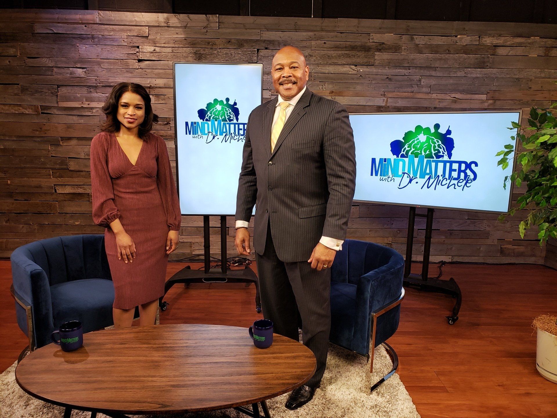 Two people stand in a studio with armchairs, a coffee table, and monitors displaying a show logo.