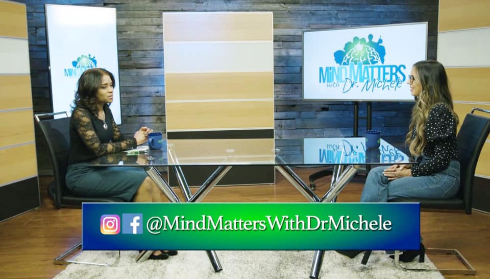 Two women converse at a glass desk in a studio with the Mind Matters With Dr Michele logo on screen.