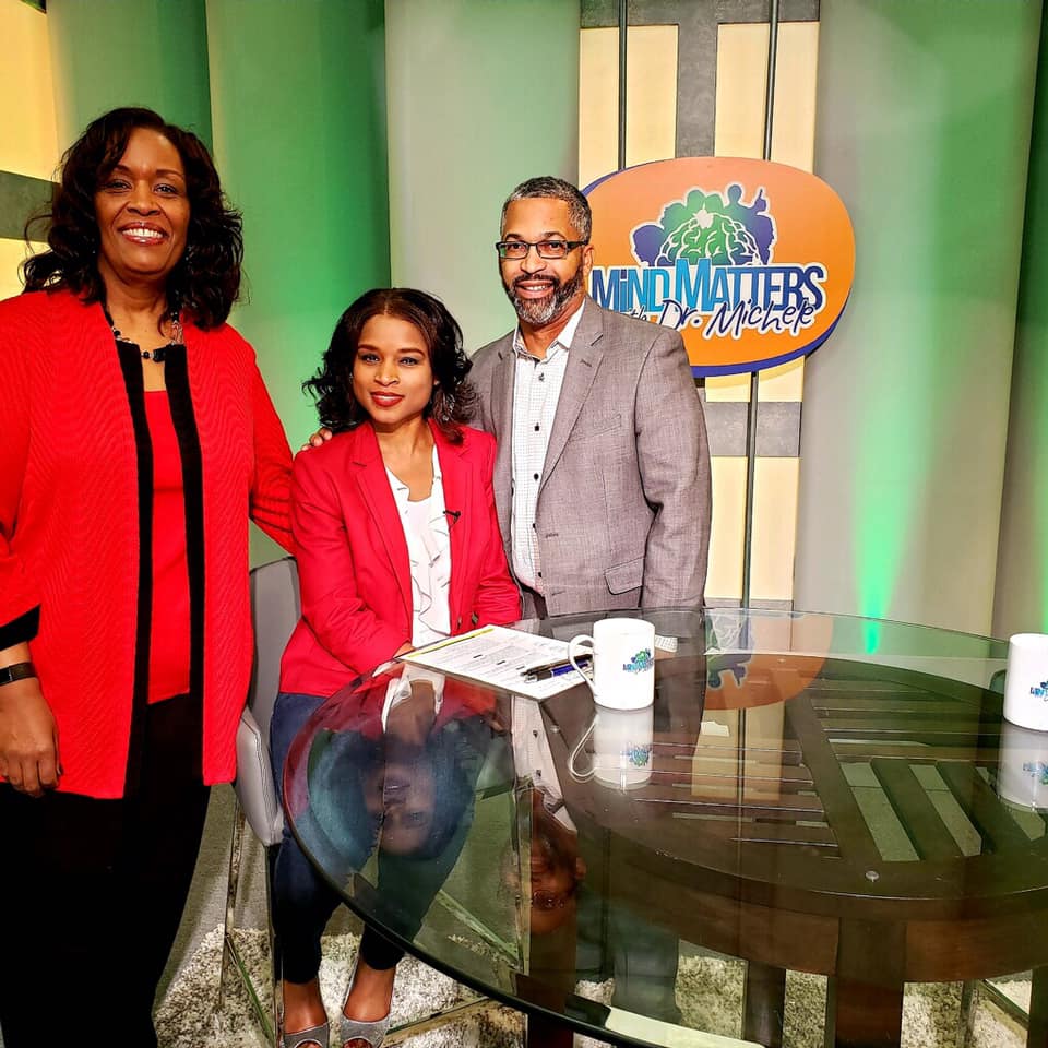 Three people standing behind a glass table in a studio beside a “Mind Matters” sign.