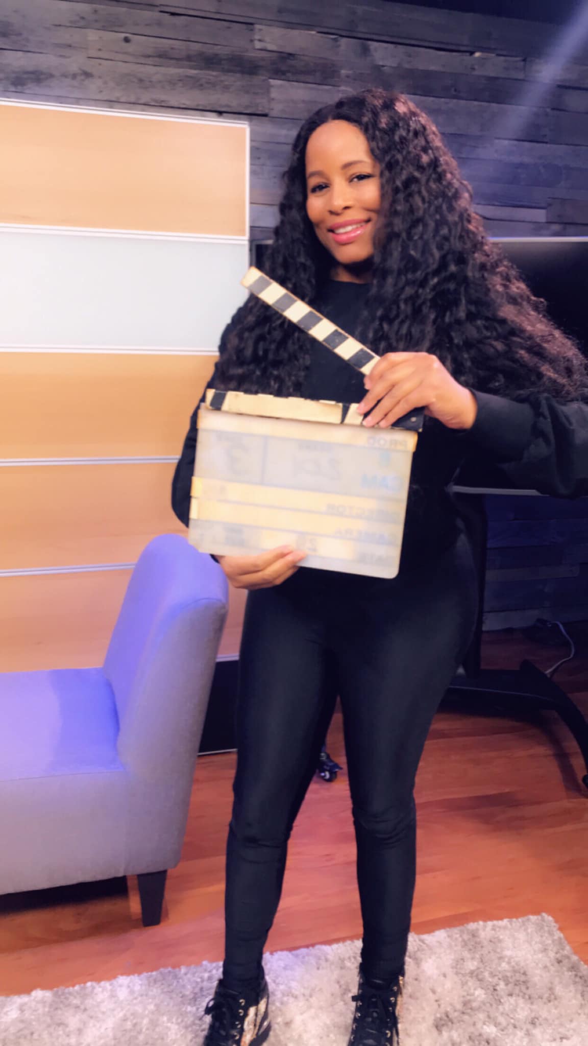 Woman in black outfit holding a cream clutch, smiling in a lounge with a purple chair and wood floor.
