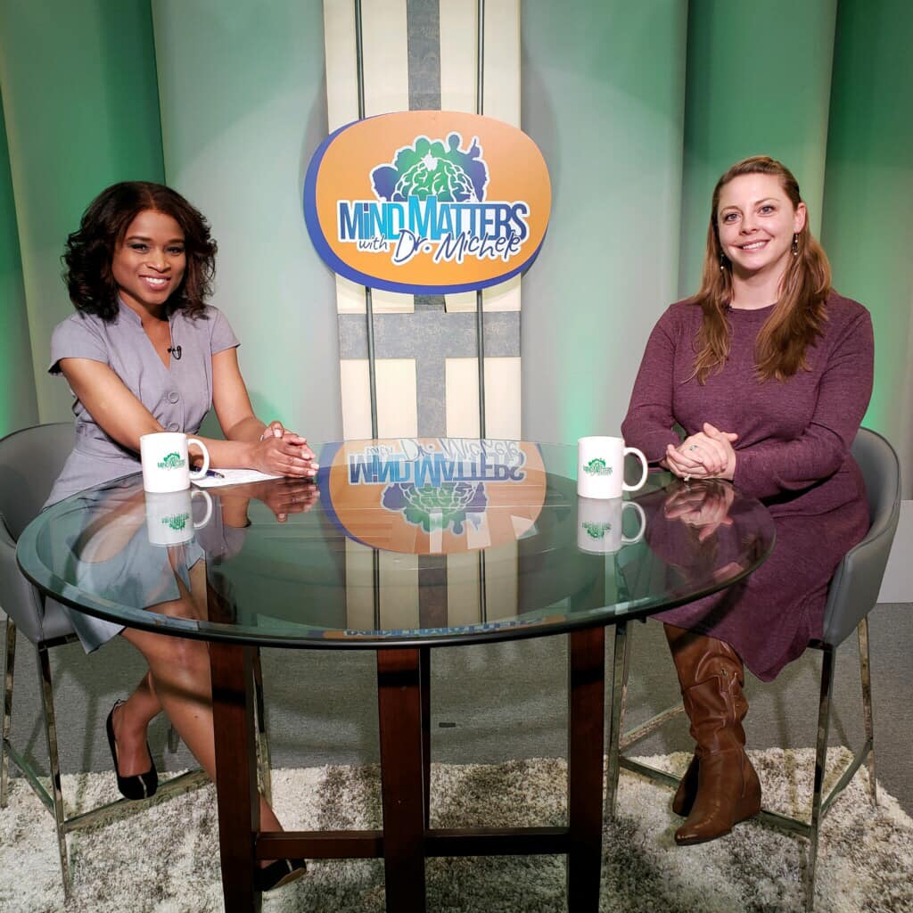 Two women seated at a glass table in a studio with mugs and a colorful logo backdrop