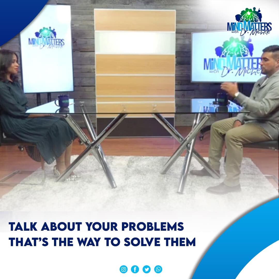 Two people seated in a studio talk show set with screens and the slogan “Talk about your problems”