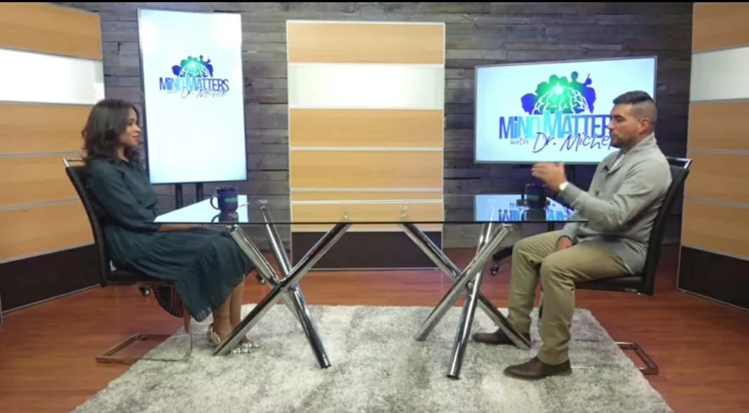 Two people seated in a studio interview setup with monitors and a table between them