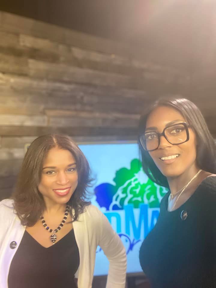 Two smiling women standing indoors in front of a TV screen and wood-paneled wall
